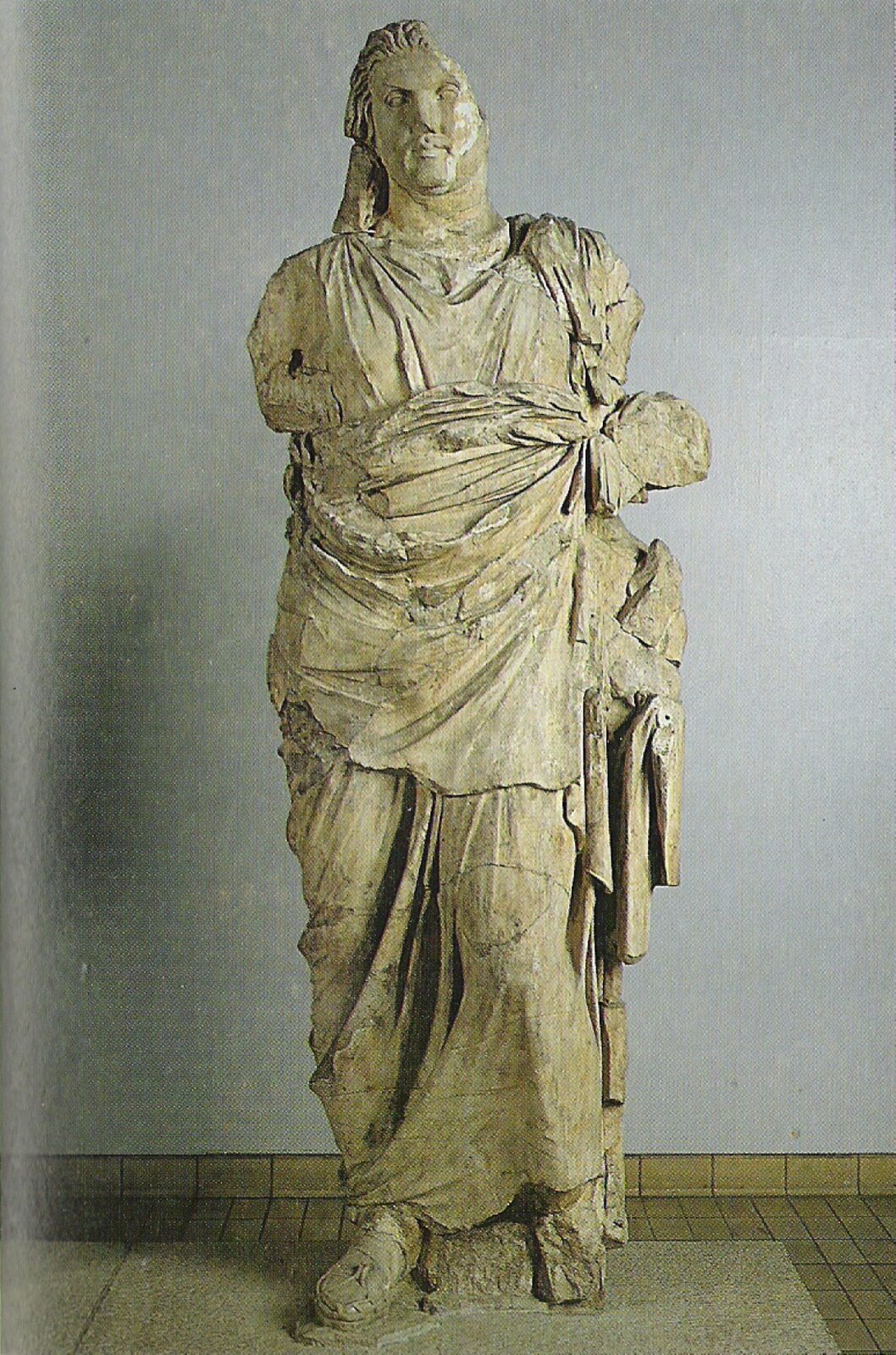 Zdjęcie przedstawia mocno zniszczoną rzeźbę mężczyzny w kolorach beżu, która znajduje się w pomieszczeniu o szarych ścianach. Fragment jego głowy odpadł, podobnie stało się z rękoma. Postać odziana jest w zwiewne szaty oraz sandały. Jego twarz jest nieco pulchna, a włosy dłuższe.