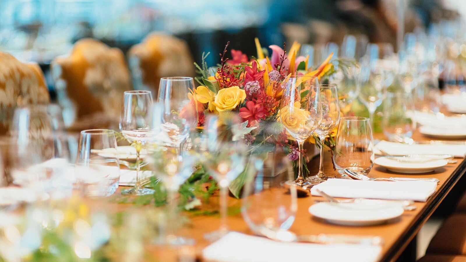 Zdjęcie przedstawia bardzo długi stół w ujęciu z ukosa, na którym widoczna jest przygotowana zastawa stołowa oraz dekoracje. Wzdłuż krawędzi stołu ustawiono nakrycia, na które składają się mały talerzyk, sztućce, serweta, dwa kieliszki do wina i niska szklanka. Pośrodku stołu widoczne są dekoracje w postaci zielonych gałązek oraz bukietu kolorowych kwiatów.