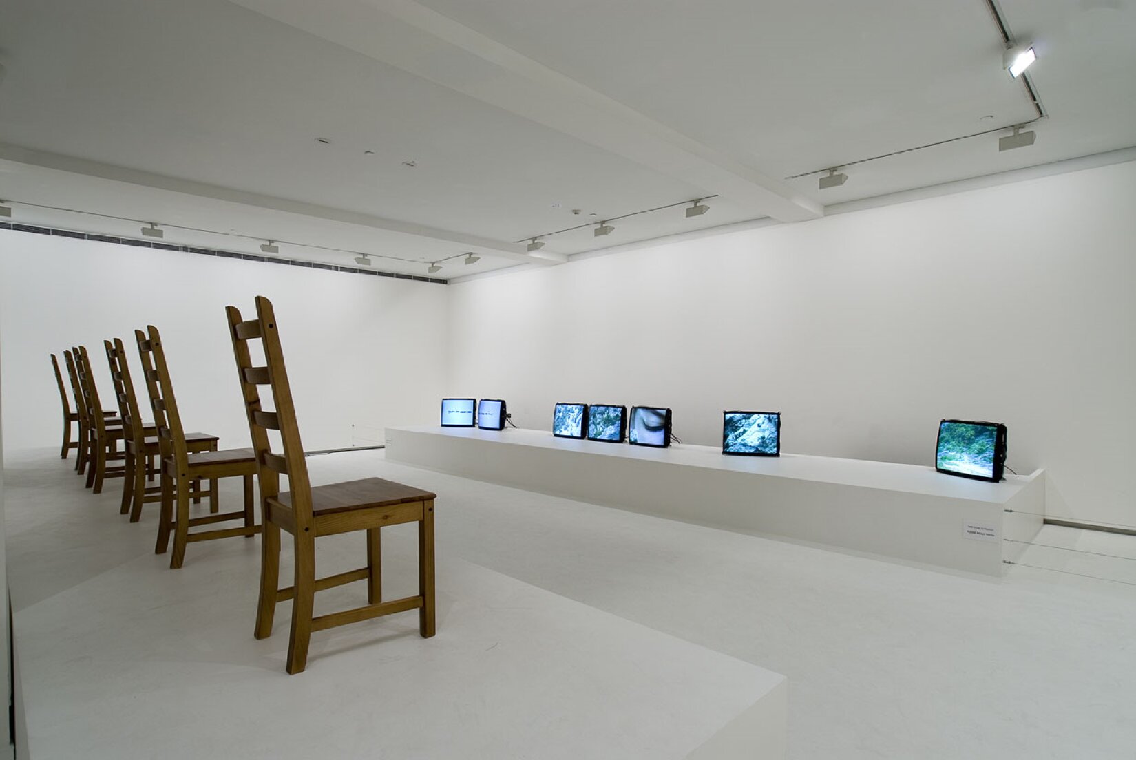 Zdjęcie przedstawia wnętrze białego pokoju, w którym znajduje się pięć drewnianych krzeseł, a przed nimi ustawione zostały niewielkie telewizory.