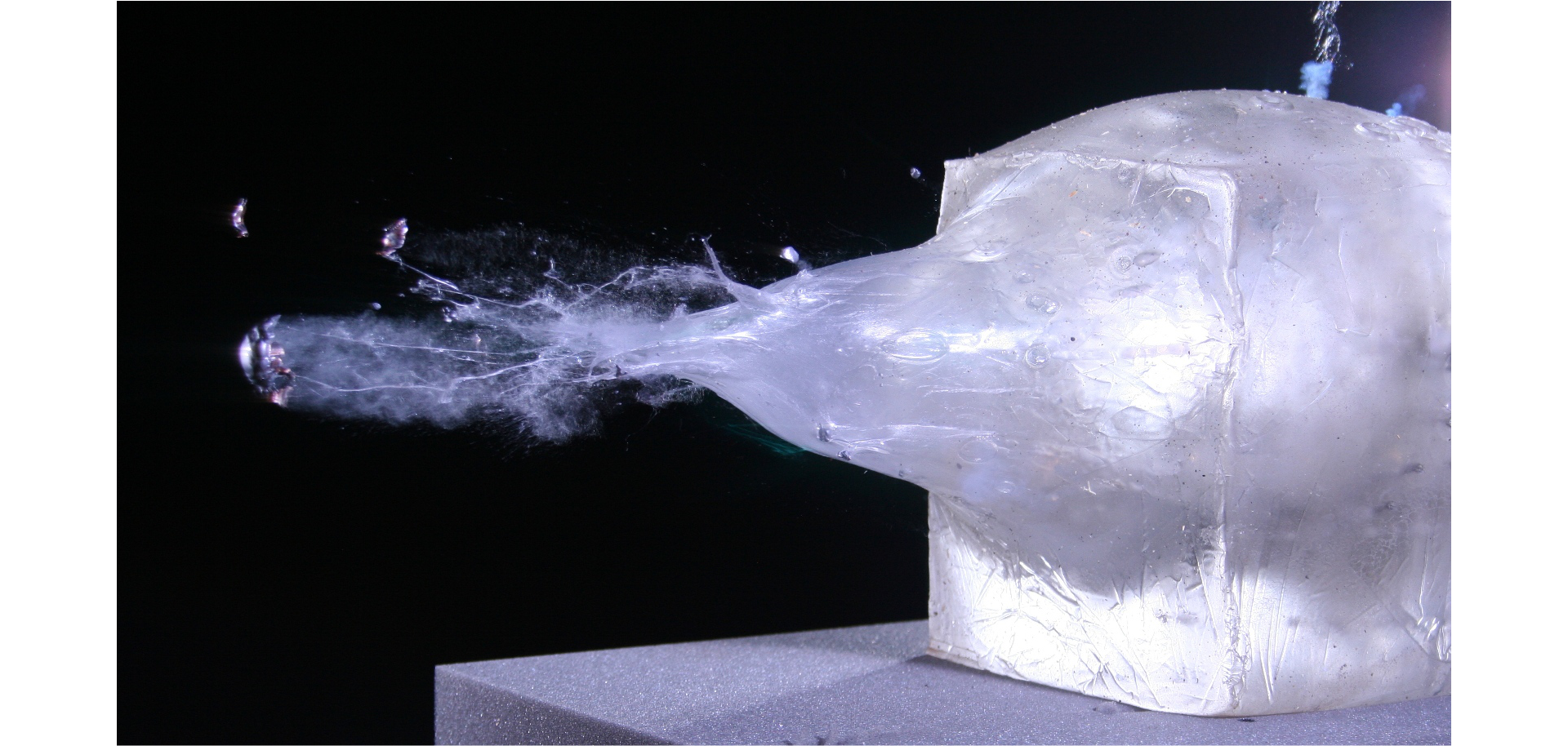 Fotografia przedstawia żel balistyczny podczas przelotu przez niego kuli. Żel ma formę dużego, półprzezroczystego prostopadłościanu. Na jego ścianie widać podłużne wybrzuszenie z pęknięciem na końcu, od którego odrywają się fragmenty substancji.