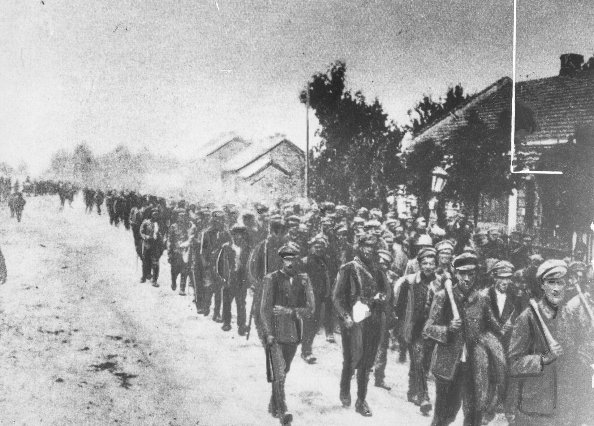 Zdjęcie przedstawia jeńców sowieckich. Są ubrani w marynarki i czapki. Idą w równych rzędach po drodze. Obok nich idzie pilnujący ich żołnierz z karabinem. Po prawej stronie są zabudowania.