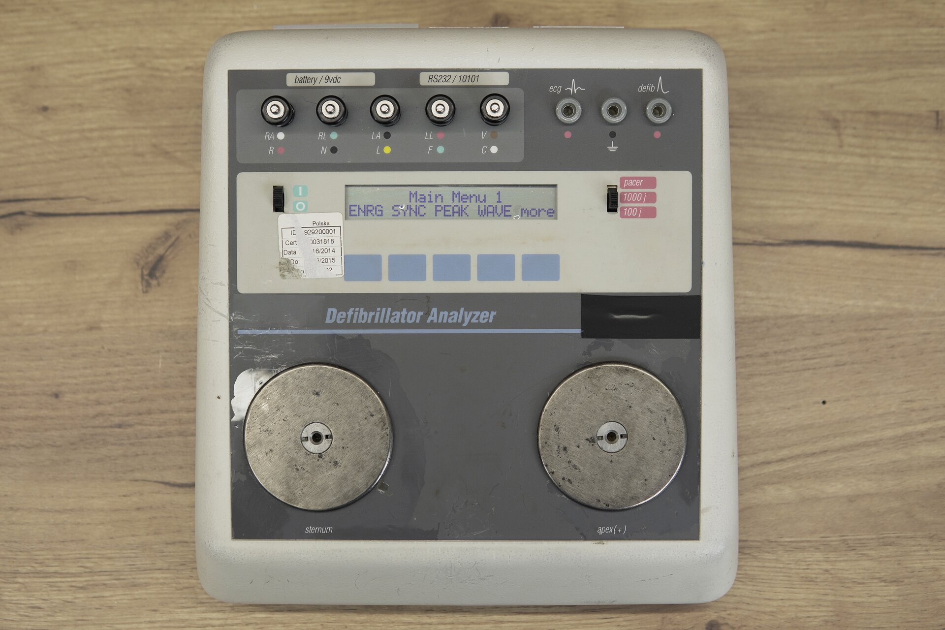 Zdjęcie przedstawia tester do defibrylatora w perspektywie od góry. Na obudowie urządzenia znajdują się dwie okrągłe, metalowe płytki, ekran, przyciski sterujące oraz rząd gniazd do podłączenia innych urządzeń. 