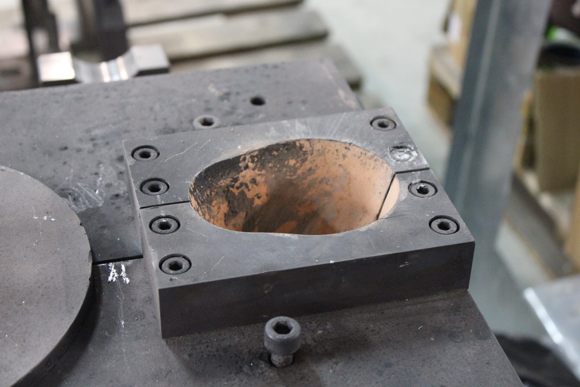 Zdjęcie przedstawia fragment zbiornika wlewowego kokili odlewniczej. Widoczna jest kostka z owalnym otworem, którym jest zbiornik wlewowy. Kostka ta przykręcona jest ośmioma śrubami do powierzchni kokili. 