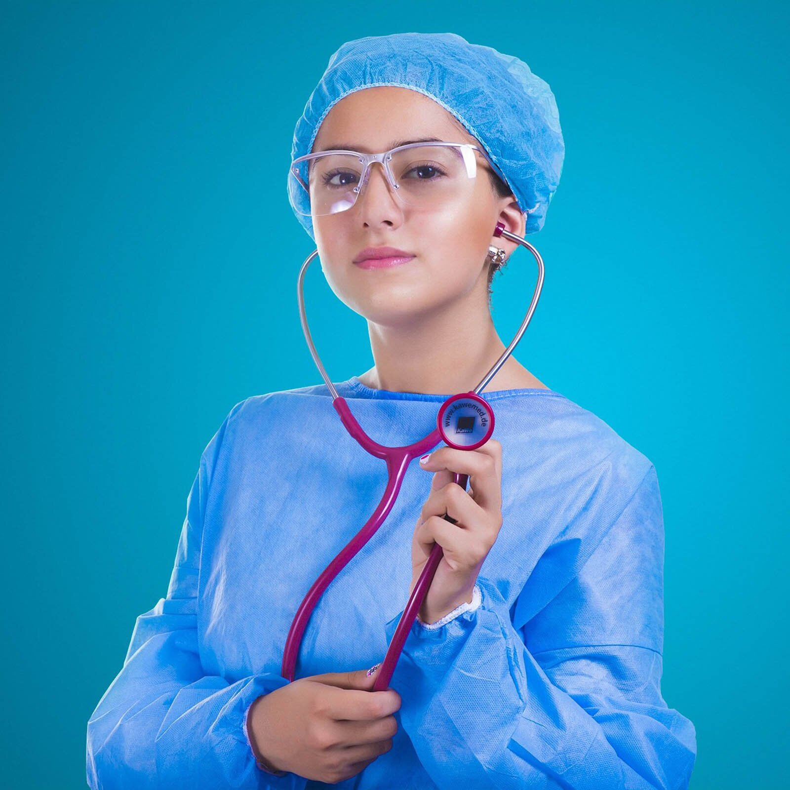 Zdjęcie przedstawia młodą kobietę ubraną w fartuch oraz czepek medyczny w kolorze niebieskim. Ponadto kobieta ma założone ochronne okulary medyczne, w uszach włożone słuchawki od stetoskopu a dolną jego część trzyma w lewej ręce, skierowanej w stronę patrzącego na zdjęcie. Transkrypcja nagrań znajduje się poniżej.