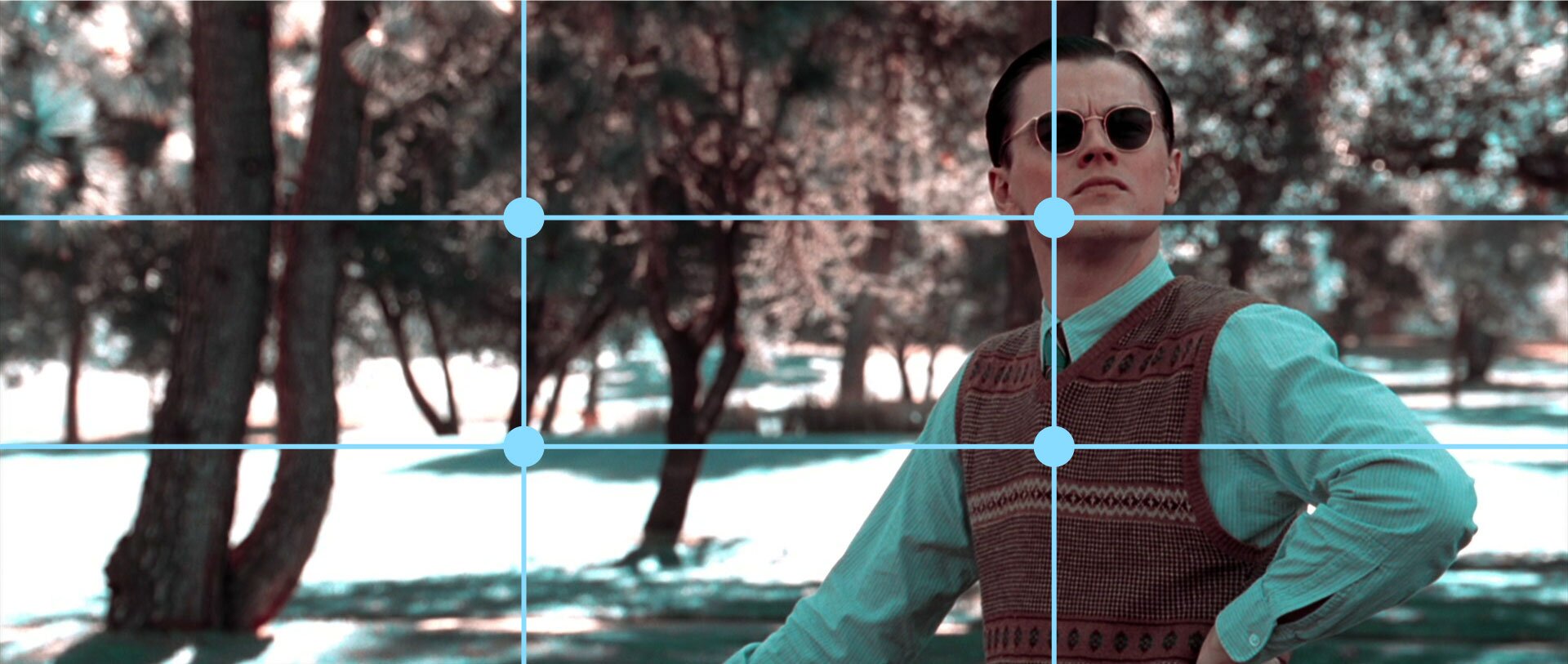 Ilustracja przedstawia Leonardo di Caprio w kadrze z filmu „Aviator”. Na ilustracji znajduje się mężczyzna ubrany w niebieską koszulę oraz wełnianą kamizelkę. W tle ilustracji znajdują się drzewa. Na ilustracji znajdują się dwie poziome oraz dwie pionowe kreski, które tworzą linie podziału kadru. Dzielą ilustrację na dziewięć równych części. Na rogach środkowego prostokąta znajdują się kropki, które wyznaczają mocne punkty kadru. W prawym górnym mocnym punkcie znajduje się twarz mężczyzny, a w dolnym środek klatki piersiowej. W lewym górnym mocnym punkcie znajdują się liście drzew, a w dolnym pień.