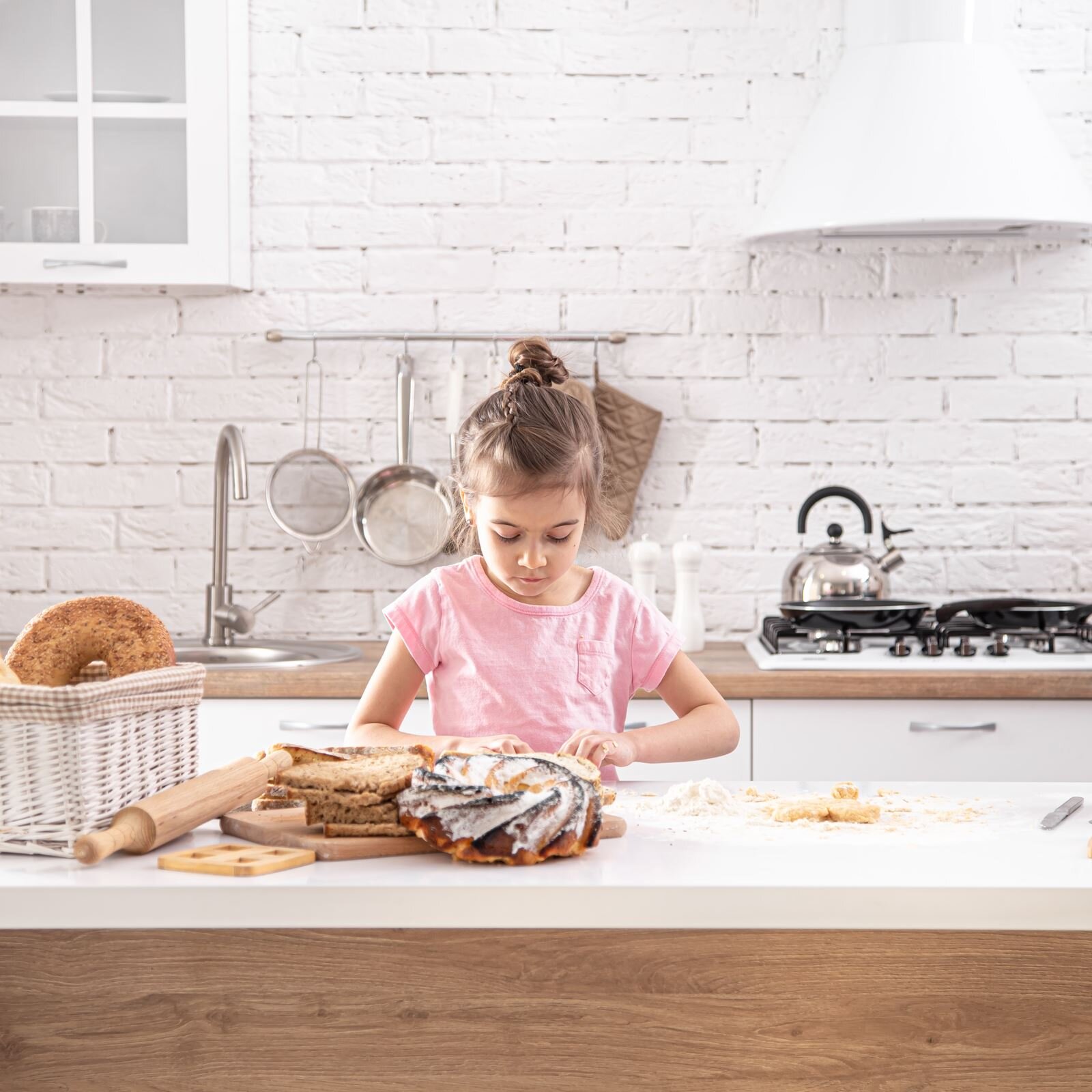 Zdjęcie przedstawia wnętrze kuchni. Na pierwszym planie widoczny jest blat, za którym stoi dziewczynka w wieku wczesnoszkolnym. Na blacie od lewej widoczne są: wiklinowy biały kosz z dużym obwarzankiem, drewniany wałek i deska, na której widoczne są pokrojone kromki ciemnego chleba oraz lukrowany wypiek w kształcie wieńca, mąka oraz przygotowane ciasto. Dziewczynka w różowej bluzce, z wysoko upiętymi włosami, ma zgięte ręce, a jej dłonie leżą na blacie. Układ dłoni wskazuje, że dziewczynka ugniata ciasto. W tle widoczne są szafki kuchenne w kolorze białym z drewnianym blatem, kuchenka gazowa ze stojącym stalowym czajnikiem i patelnią oraz zlew. Na przymocowanym do ściany drążku wiszą garnek, sitko i rękawica kuchenna.