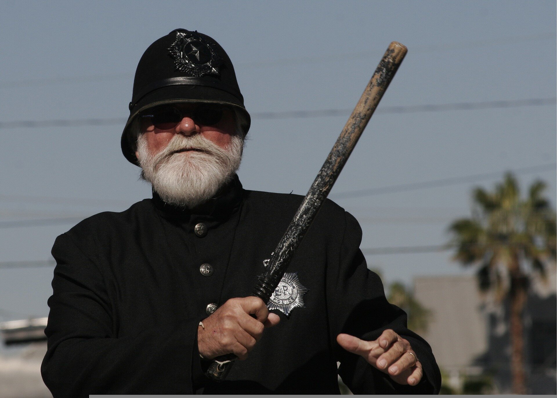 Zdjęcie przedstawia mężczyznę w stroju brytyjskiego policjanta z pałką w ręku. Na jego lewej piersi widoczna srebrna odznaka. 