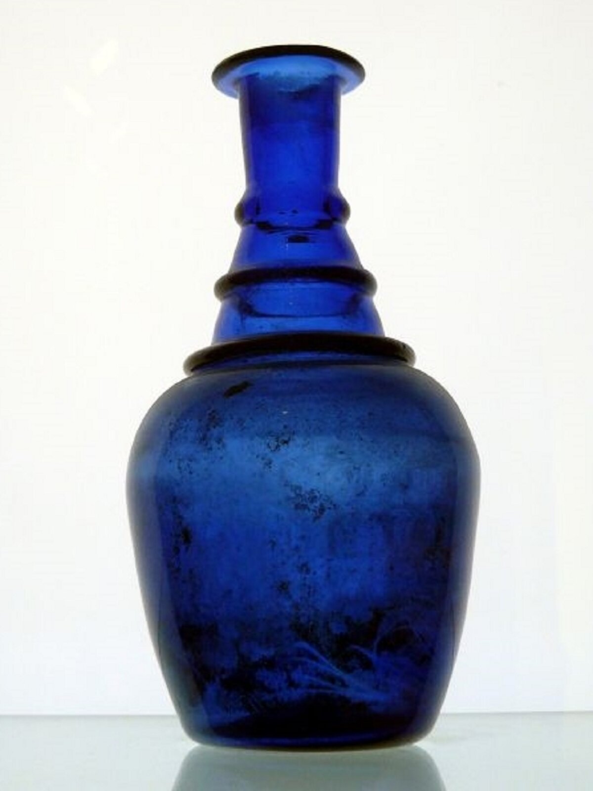 Grafika przedstawia naczynie szklane zabarwione związkami kobaltu(dwa) lub miedzi(dwa) z wąską szyjką w kształcie wazonu. Naczynie jest intensywnie niebieskie. Kolor niebieski ma miejscami czarne plamy.