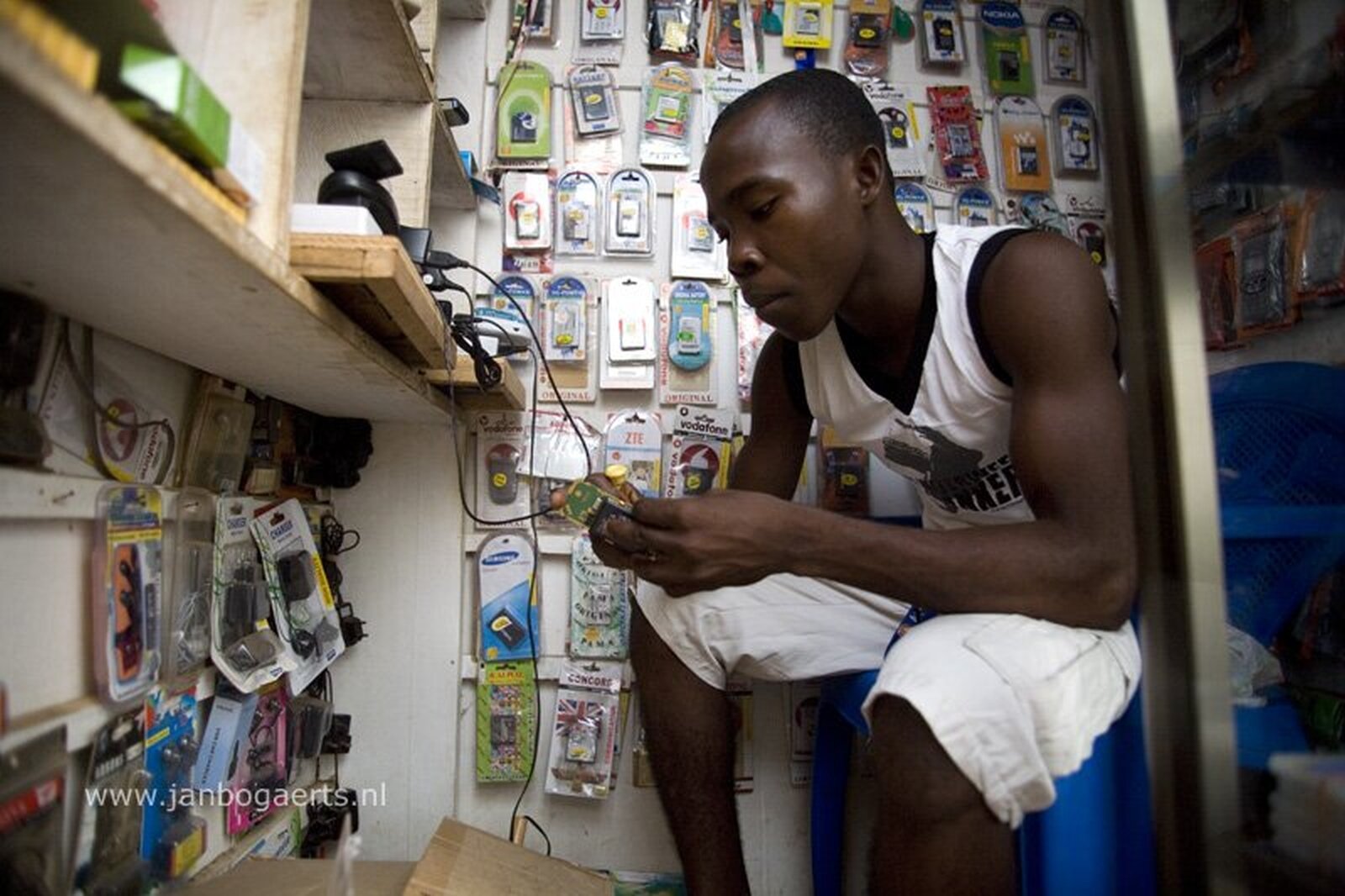 Zdjęcie przedstawia niewielkie pomieszczenie, w którym na krześle siedzi czarnoskóry mężczyzna ubrany w krótkie spodenki i bezrękawnik. Na ścianach pomieszczenia zawieszona duża ilość akcesoriów i części do telefonów, na przykład baterii.