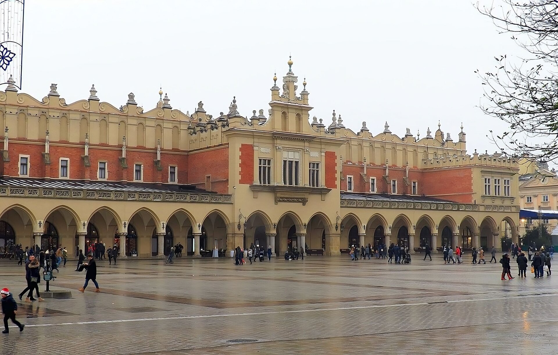 Na zdjęciu widok współczesny na ścianę Sukiennic w Krakowie z arkadami w części parterowej na całej długości ściany, pokrytych dachem. W połowie konstrukcji bocznej ściany, zarówno w parterowej części nad kolumnadą i w górnej kondygnacji widoczny wykusz z prostokątnymi oknami. Ściana na poziomie powyżej kolumnady wykonana z czerwonej cegły. Szczyt ściany budynku zdobi renesansowa attyka, z widocznymi wolutami i sterczynami na szczytach attyki osłaniającej dach. Na pierwszym planie fragmenty rynku w Krakowie, zdjęcie wykonane w deszczowy dzień.