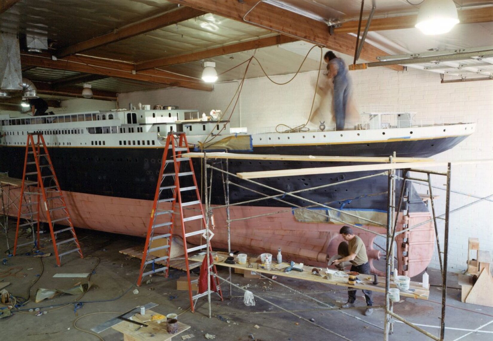 Ilustracja przedstawia prace wykonywane przy modelu Titanica w skali 1:20. Na ilustracji znajduje się wielki kadłub statku w kolorach jasnoczerwonym, czarnym i białym. Z prawej strony ilustracji stoi rusztowanie, na którym są deski leżące na dwóch poziomach. Na deskach, które są niżej jest postawionych wiele przedmiotów. Za rusztowaniem stoi mężczyzna o krótkich, czarnych włosach. Ma na sobie brązową koszulkę z krótkim rękawem, ciemne, długie spodnie i brązowe buty. Po lewej stronie obrazka i na środku stoją 2 wysokie drabiny w kolorze pomarańczowo‑srebrnym. Przed drabiną, która jest na środku ilustracji, stoi drewniany, kwadratowy stół. Są na nim różne przedmioty. Na górze statku znajduje się postać, która ma ciemne włosy. Ubrana jest w jasną bluzkę z krótkim rękawem i niebieskie ogrodniczki. Kadłub statku znajduje się w wysokim pomieszczeniu. Podłoga jest w kolorze szarym, leży na niej dużo przedmiotów. Sufit jest w kremowym kolorze z brązowymi belkami, które są ułożone wzdłuż i w poprzek. Z sufitu zwisają 3 duże lampy. 2 z nich są włączone. Za kadłubem statku znajduje się kremowa ściana.
