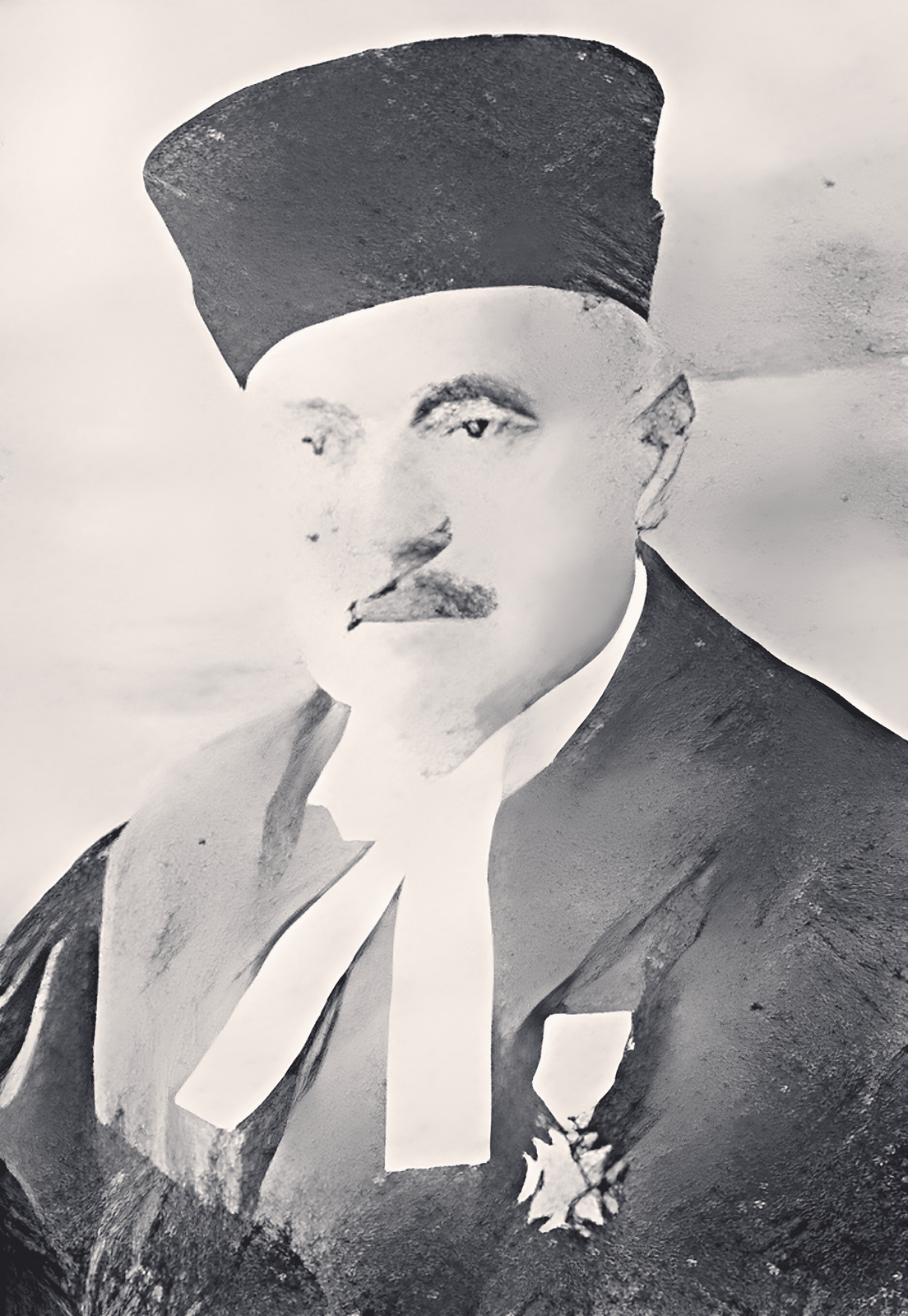 Zdjęcie przedstawia starszego mężczyznę, który ma czarne brwi i mały, cienki wąs. Na głowie ma wysoką, czarną czapkę. Ubrany jest w czarną szatę, spod której wystaje biała wstążka. Na piersi ma przypiętą odznakę w kształcie gwiazdy. Tło jest jednolite. Zdjęcie jest czarno‑białe.