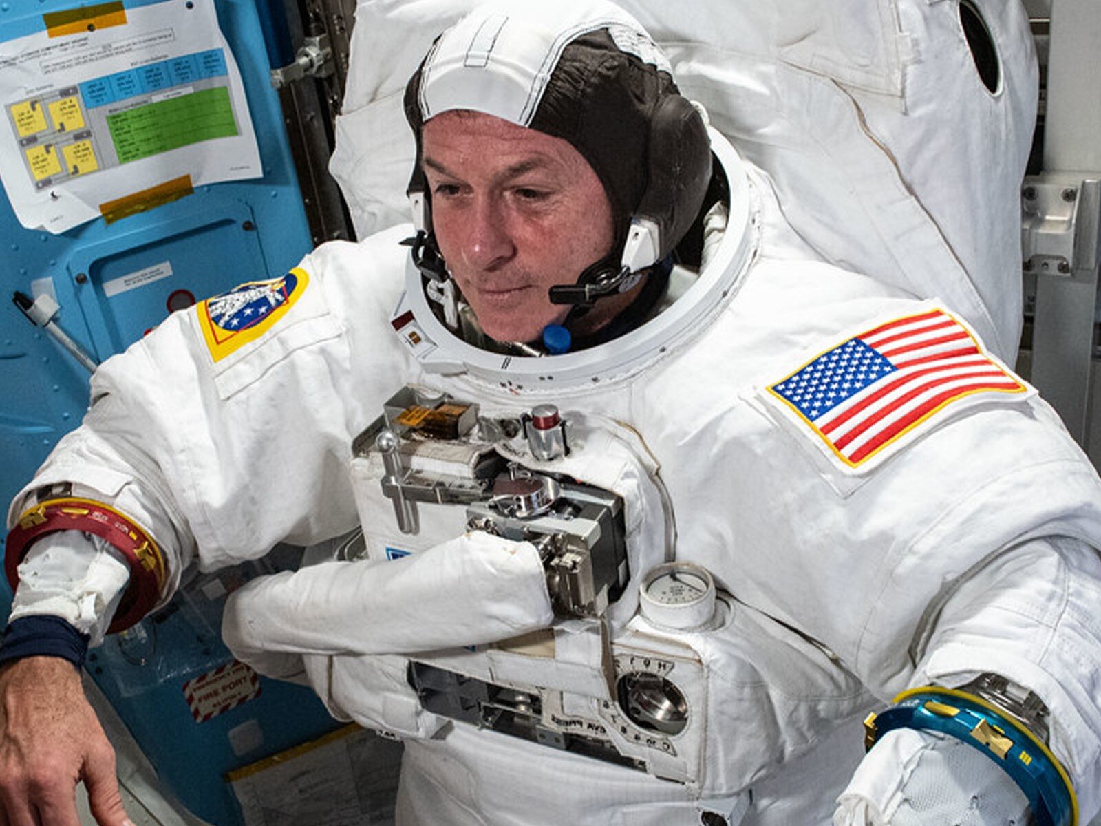 Zdjęcie przedstawia siedzącego mężczyznę, ubranego w biały skafander astronauty, czarno‑białe nakrycie głowy, przylegające do głowy i wyposażone w słuchawki i mikrofon. Na skafandrze, na wysokości klatki piersiowej, widoczne jest mocowanie do plecaka kosmonauty z tlenem, do którego prowadzi biała rura i który znajduje się z tyłu. Na ramionach skafandra widoczne są naszywki. Na lewym ramieniu widoczna jest naszywka z flagą amerykańską, a na prawym żółto‑niebieska naszywka z rakietą. 