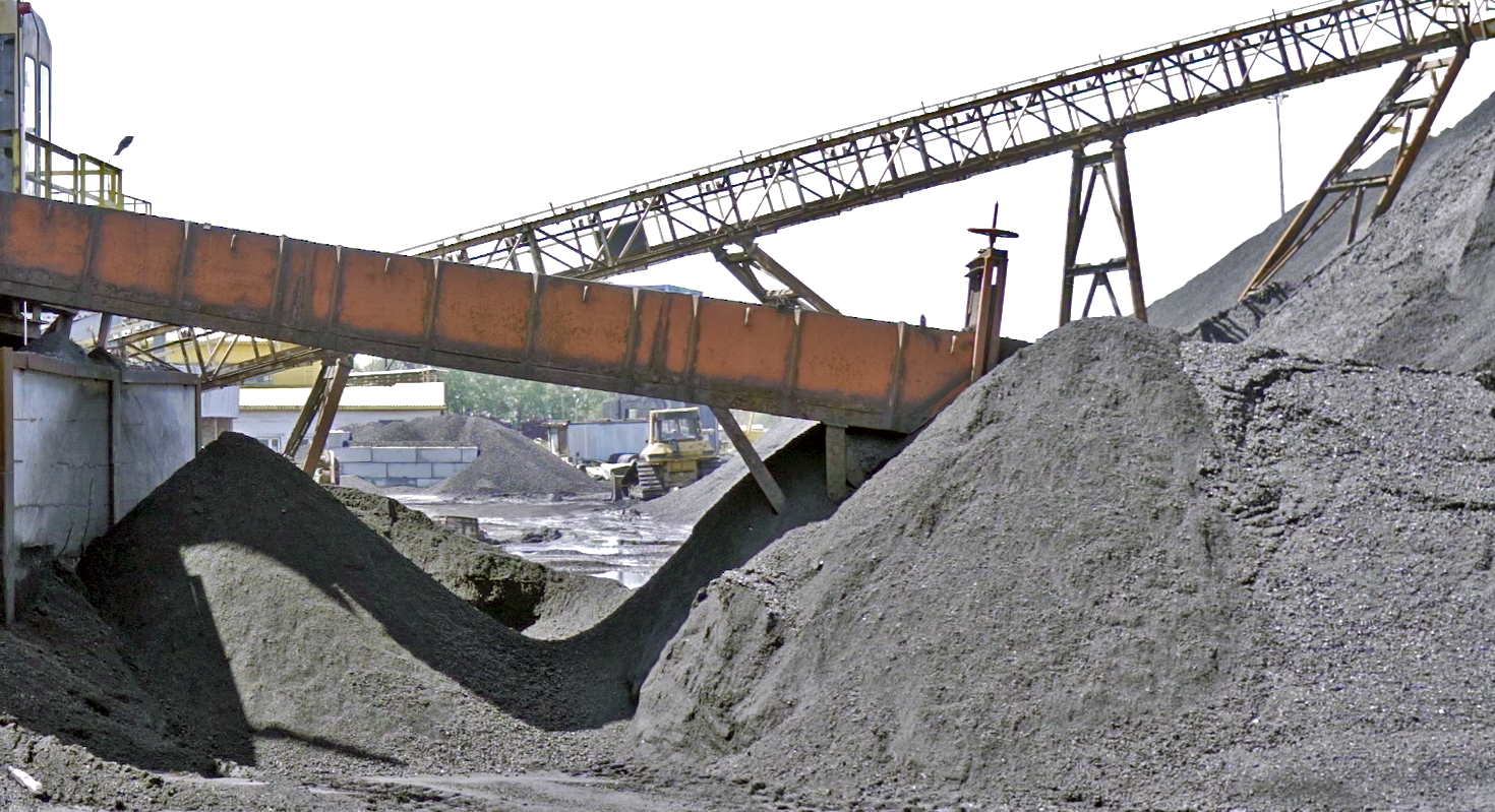 Na zdjęciu widać zwałowisko węgla. Węgiel jest składowany w formie pryzmy. Widoczny węgiel ma małe rozmiary ziarna. W tle widać przenośniki. Przenośniki są nachylone względem gruntu. Oba przenośniki stanowią konstrukcję kratową modułową. Oba są zbliżone kształtem do rynny.Poniżej zdjęcia widoczny jest przycisk multimedialny z nagraniem audio. Treść nagrania jest zgodna z tytułem zdjęcia.