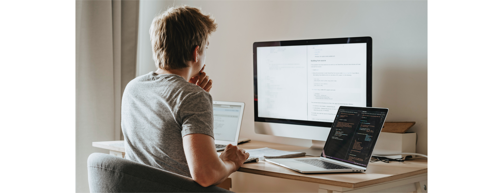 Zdjęcie przedstawia młodego chłopaka siedzącego w fotelu przy biurku. Chłopak siedzi tyłem do kadru. Na biurku, w centralnej części znajduje się duży monitor. Obok chłopaka, po lewej i po prawej stronie na biurku znajdują się dwa otwarte laptopy.