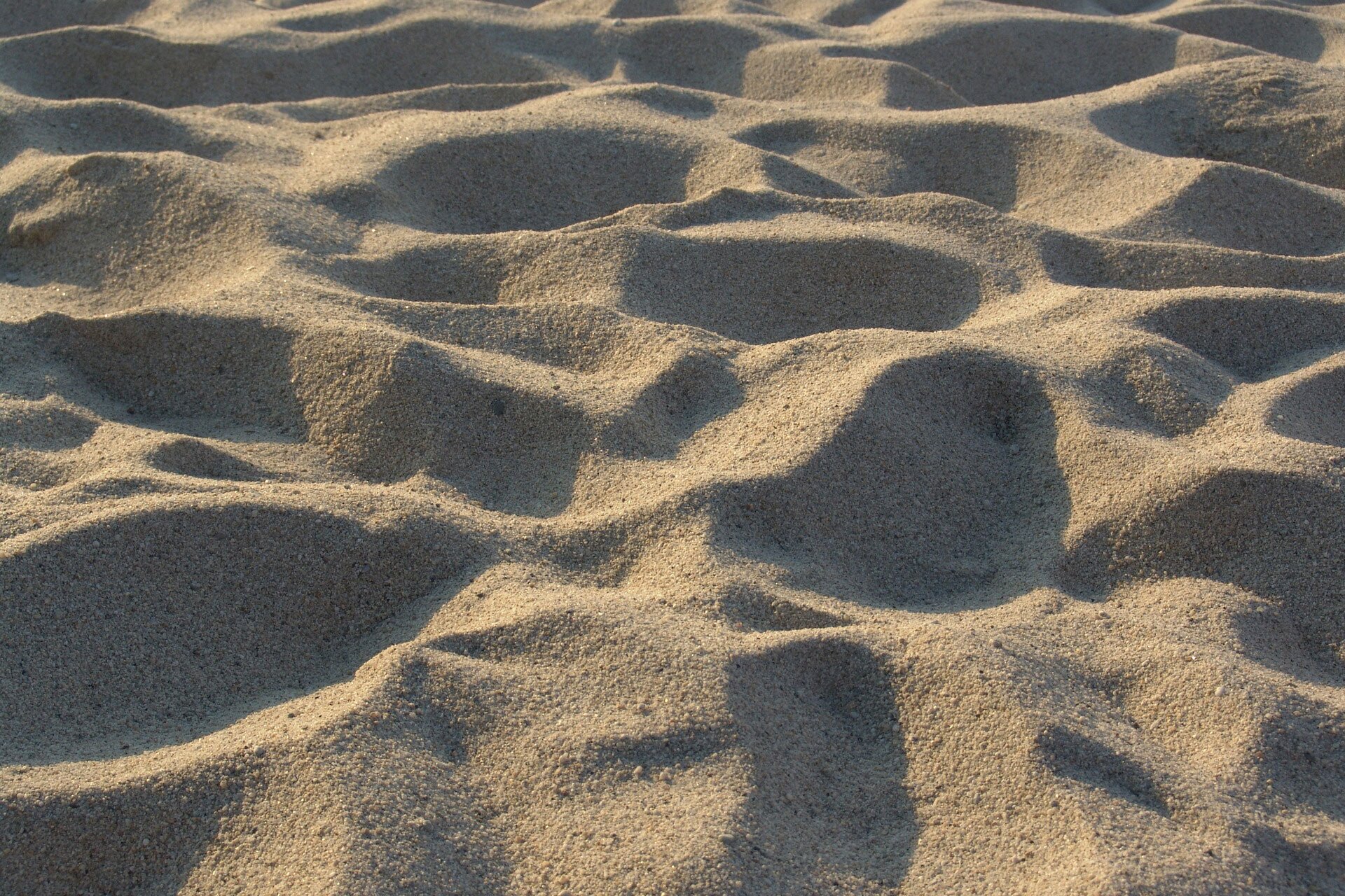 Fotografia prezentuje sypki piasek. Fotografia przedstawia piasek w naturze na plaży z licznymi nierównościami stworzonymi przez wiatr lub człowieka.
