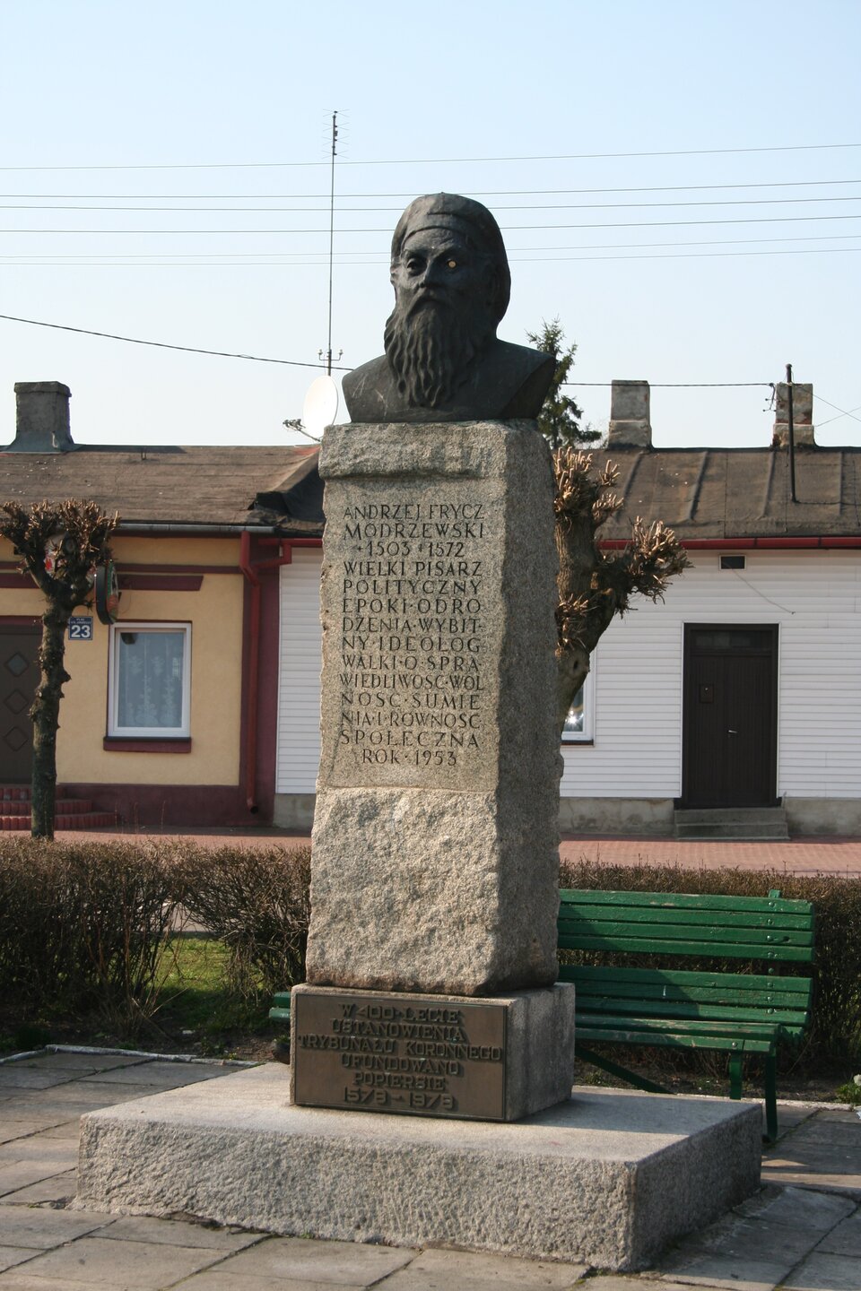 Zdjęcie przedstawia pomnik, który składa się z prostokątnego obelisku na planie kwadratu, zakończonego popiersiem starszego mężczyzny z długą brodą. Mężczyna ma owalną twarz, wydatny nos. Nosi długą, gęstą brodę i wąsy. Na głowie ma płaską czapkę. Na obelisku widnieje napis: Andrzej Frycz‑Modrzewski 1503‑1572. Wielki pisarz polityczny epoki odrodzenia. Wybitny ideolog walki o sprawiedliwość, wolność sumienia i równość społeczną. Rok 1953. Obelisk stoi na szerokiej podstawie i prostokątnym, grubym kamieniu, nieco większym niż pomnik. Na nim znajduje się napis: W 400‑lecie ustanowienia Trybunału Koronnego ufundowano popiersie 1578 -1978. W tle znajdują się parterowe, niewielkie domki, tuż za obelskiem stoi zielona, parkowa ławka, za ławką żywopłot.