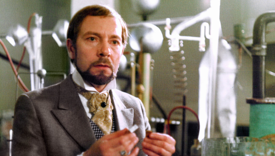 Zdjęcie przedstawia aktora, Jerzego Kamasa, grającego rolę Wokulskiego. Mężczyzna jest ubrany w surdut, kamizelkę i krawat. Znajduje się w laboratorium chemicznym.