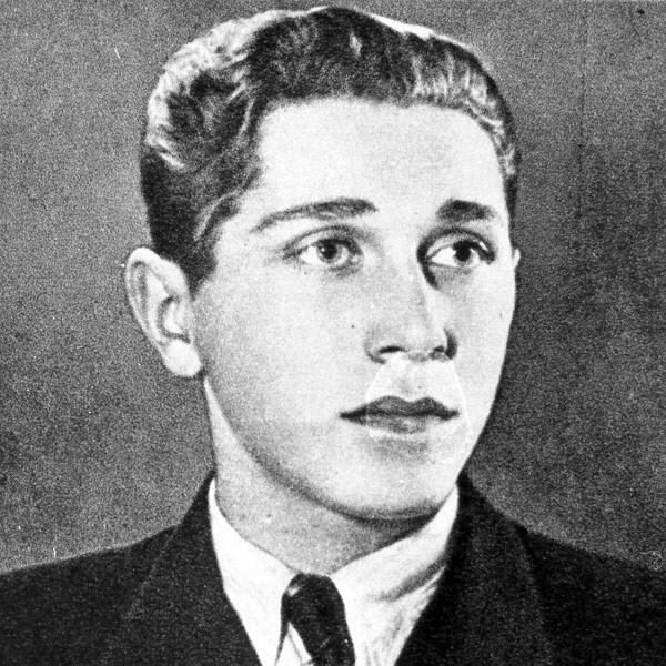 Fotografia przedstawia portret młodego mężczyzny z ciemnymi włosami zaczesanymi do tyłu, jest ubrany w garnitur, koszulę i krawat. Patrzy przed siebie.