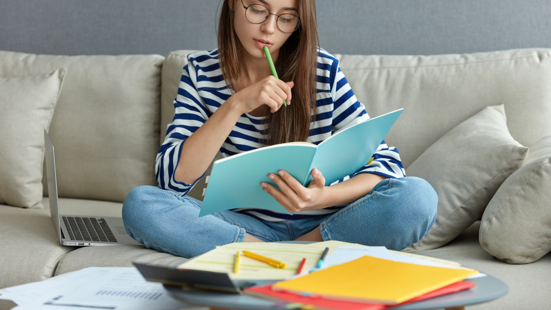 Zdjęcie przedstawia młodą dziewczynę z długimi włosami w okularach. Dziewczyna siedzi po turecku na kanapie. W ręku trzyma otwarty zeszyt i spogląda w niego. Drugą ręką przykłada do twarzy coś do pisania. Obok niej na kanapie leży otwarty laptop. Przed dziewczyną na stoliku leżą różne notatki.