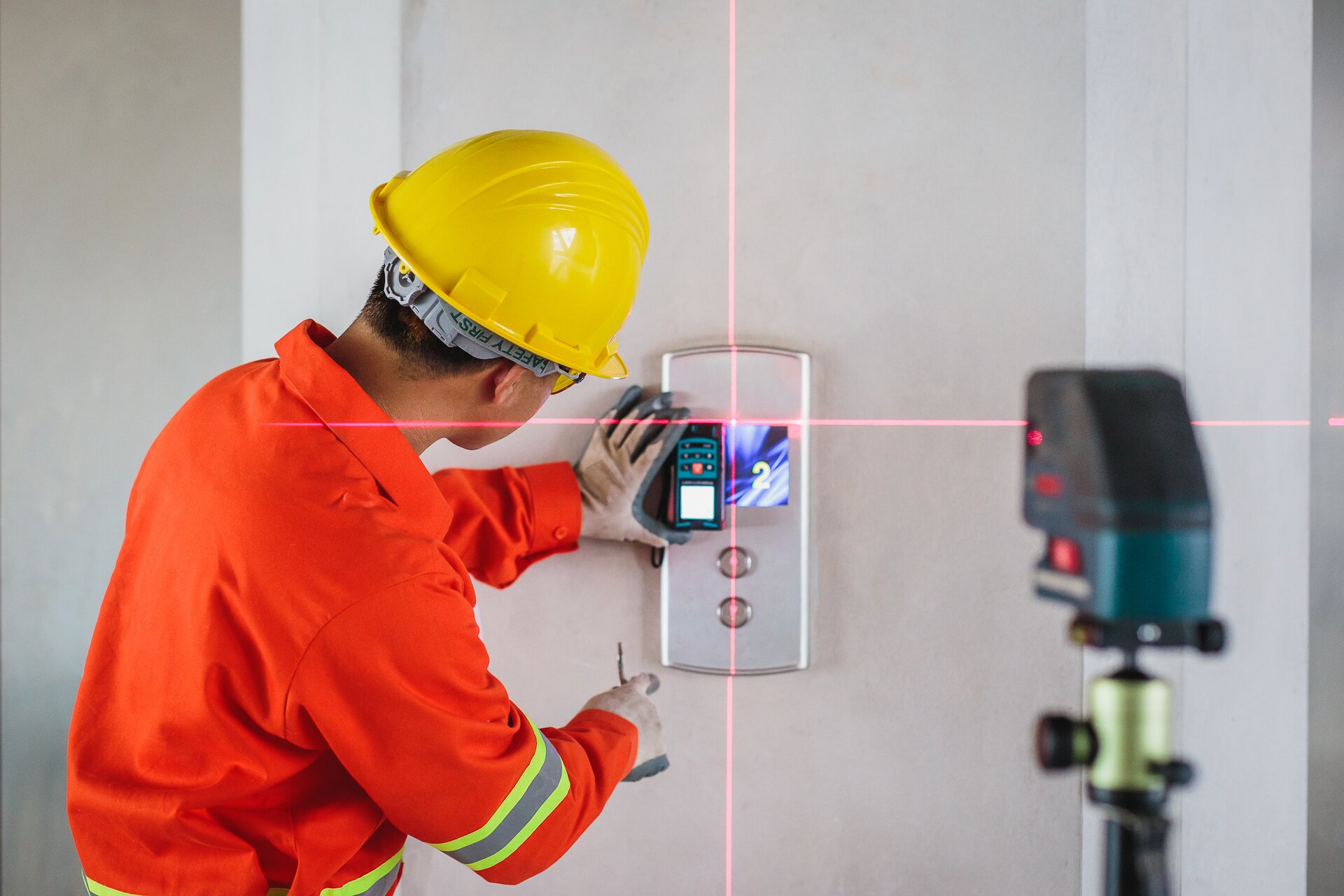 Zdjęcie przedstawia poziomicę laserową w użyciu. Urządzenie stoi na stojaku i emituje linie laserowe, które tworzą na ścianie krzyż.