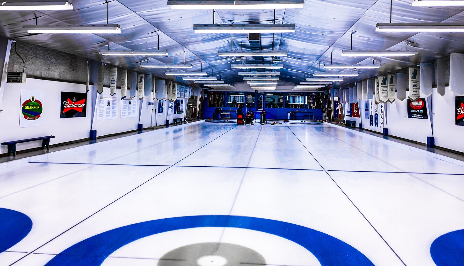 Zdjęcie przedstawia lodowisko, na którym znajdują się trzy tory do gry w curling. Każdy tor jest podłużny i biały, zakończony trzykolorowym kołem. 