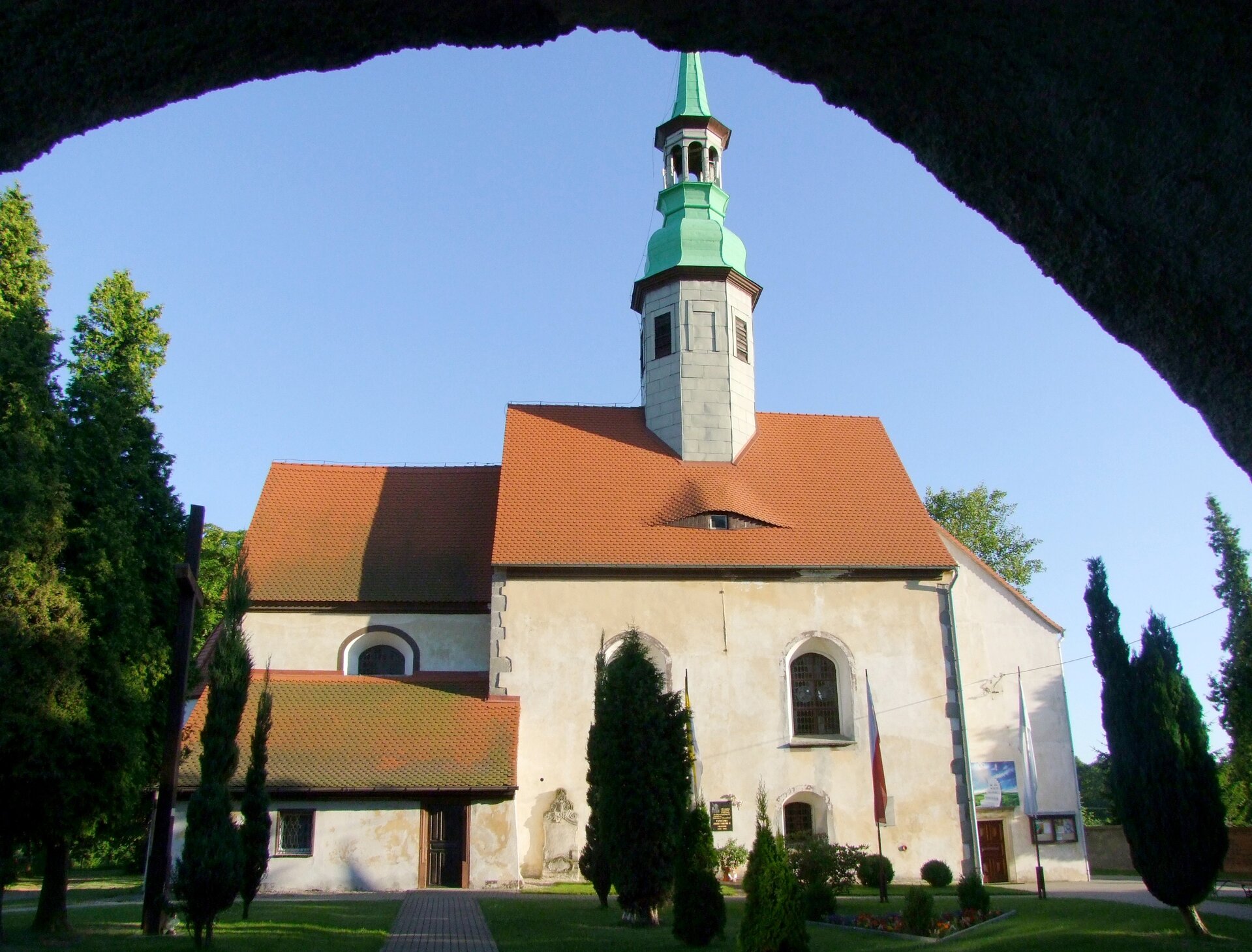 Na zdjęciu znajduje się mały kamienny kościół o stromym czerwonym dachu i wysokiej wierzy z ostrym zielonym zadaszeniem. Wokół kościoła znajduje się park z drzewami. 