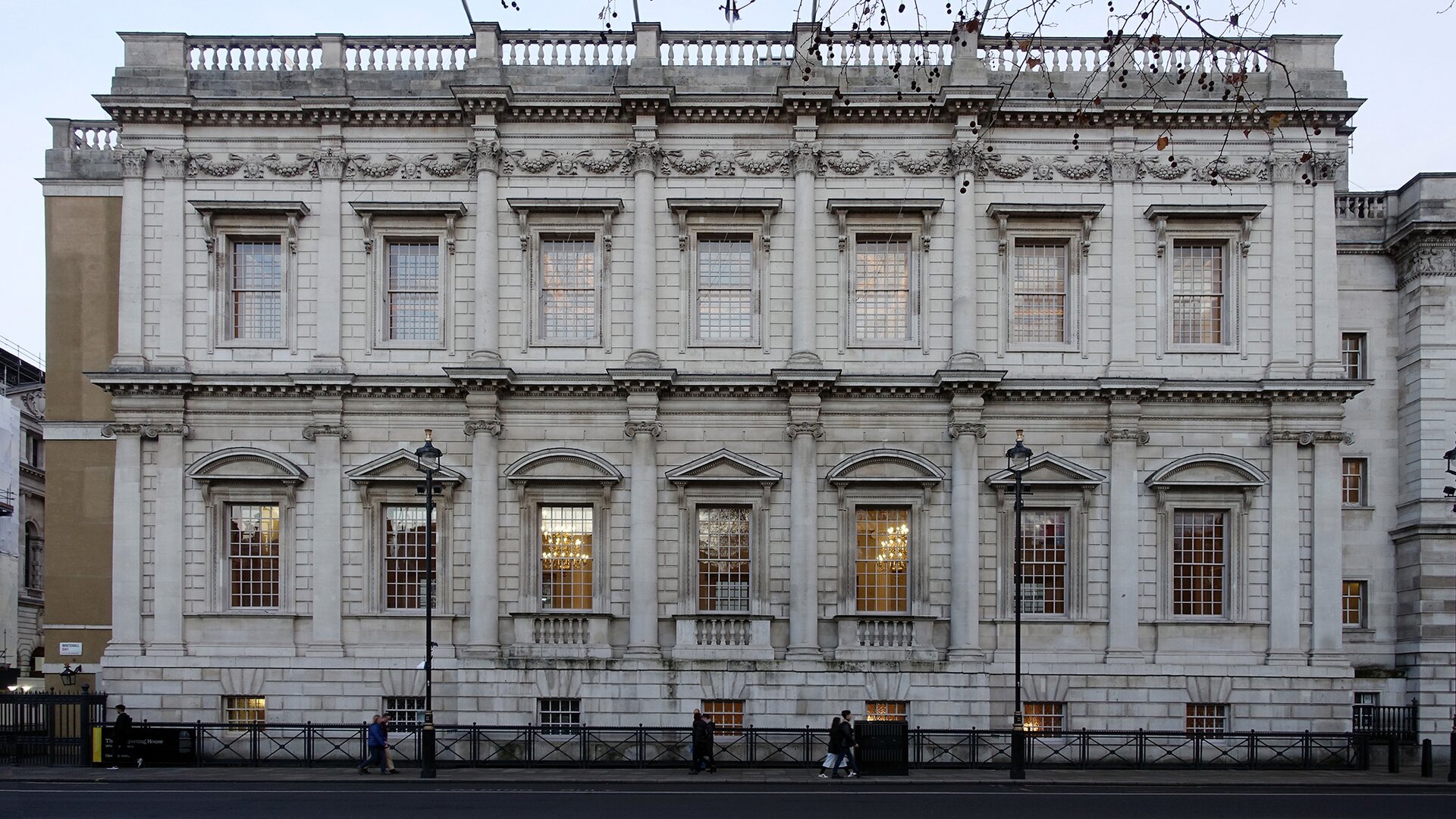 Zdjęcie przedstawia Banqueting House w Londynie, fotografię wykonał Steven Zucker. Na fotografii przedstawiono prostokątny budynek, wykonany jest z szarej cegły. Dach jest płaski, a linia dachu jest określona przez balustradę. Na fasadzie znajdują się kolumny, które dzielą okna. Budynek ma trzy piętra. Najniższe z pięter ma małe, kwadratowe okna. Następne dwa piętra mają większe, prostokątne okna. Środkowe okna zwieńczone są naprzemiennie trójkątnymi i segmentowymi frontonami, natomiast górne są nieozdobione. Przez okna budynku widać ozdobne, złote żyrandole. Przed budynkiem znajduje się ulica oraz chodnik, po którym spacerują przechodnie.