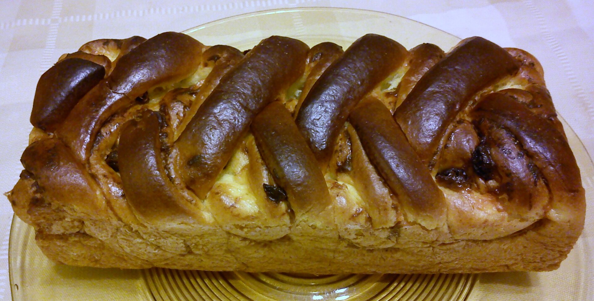 Na zdjęciu znajduje się wypiek o błyszczącej skórce w kolorze brązowym, spleciony w warkocz. Górna część jest bardziej przypieczona niż dolna. Ciasto leży na szklanym, dużym talerzu w prążki. W tle widoczny jest biały obrus.