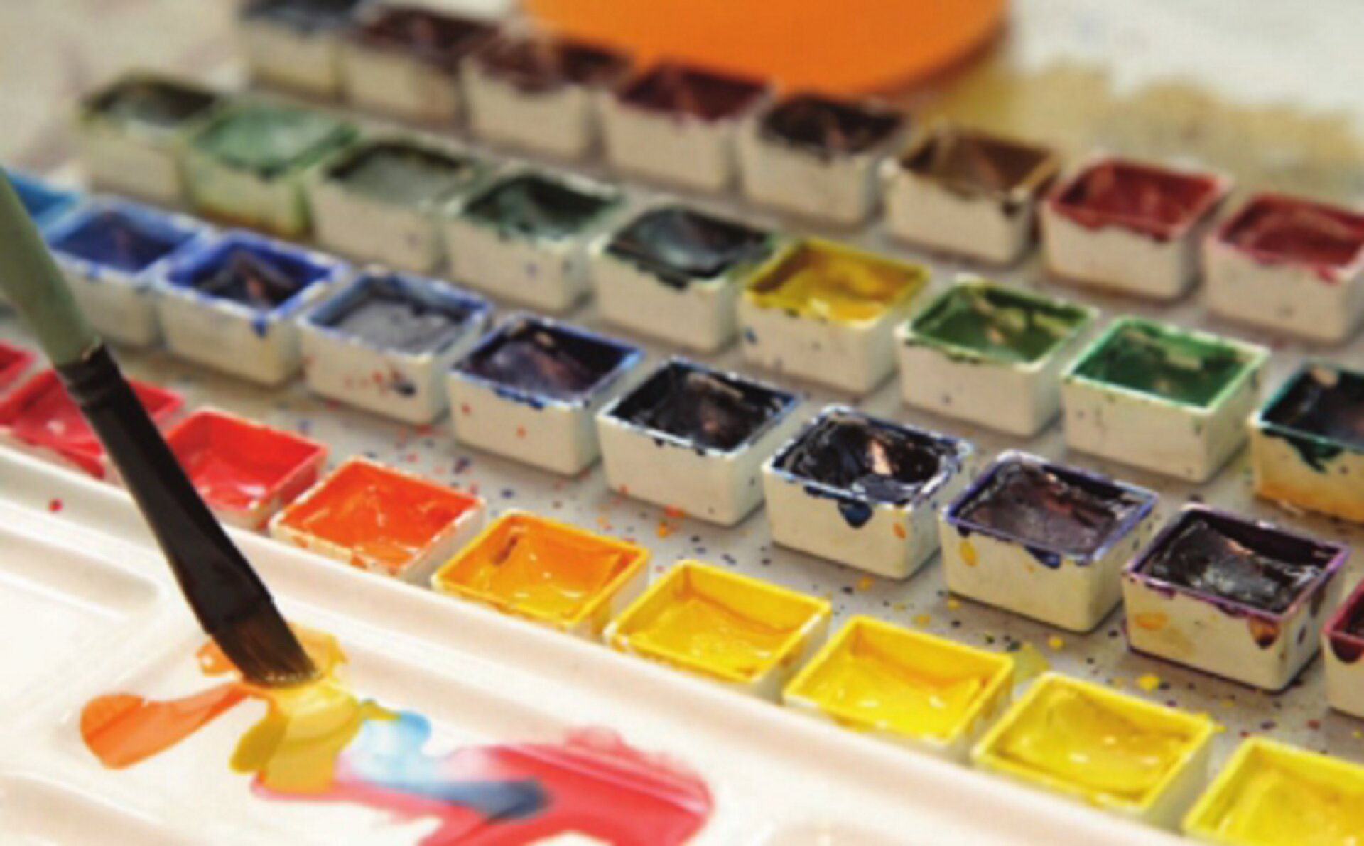 Ilustracja przedstawia farby akwarelowe w kostkach. Zdjęcie ukazuje malutkie, kwadratowe pojemniczki, w których znajdują się różne kolory i odcienie farb. Z lewej strony ilustracji znajduje się pędzel mieszający różne kolory.