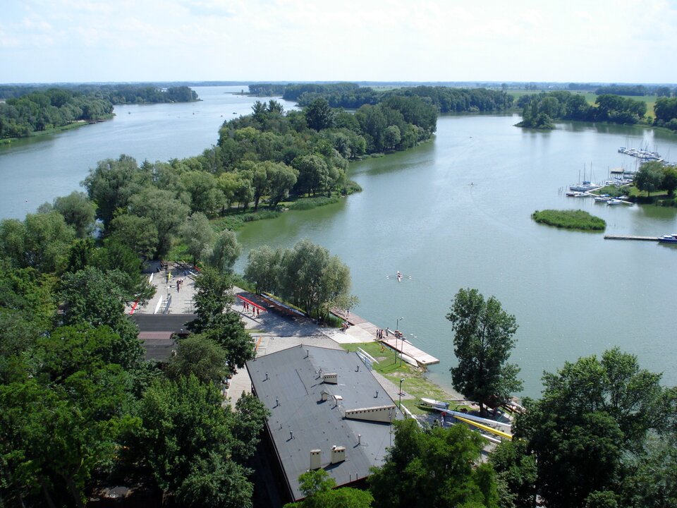 Na zdjęciu rozległy teren pokryty wodą aż po horyzont, przez który wiodą pasy lądy pokryte drzewami. U dołu zdjęcia na szerszym fragmencie lądu znajdują się zabudowania - widoczny jest duży szary dach , pomost wzdłuż brzegu.