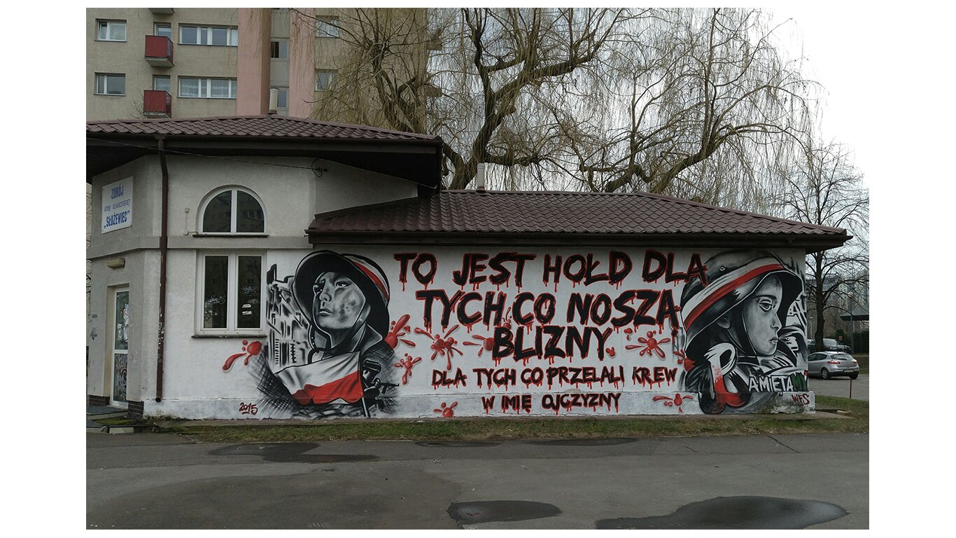 Fotografia barwna przedstawiająca jeden z wielu murali poświęconych powstaniu warszawskiemu. Po obu stronach portrety młodych powstańców w hełmach z biało-czerwoną opaską. Po lewej stronie biało-czerwona flaga, po prawej stronie biało-czerwony symbol Polski Walczącej - litera P zakończona kotwicą. W środku napis: To jest hołd dla tych, co noszą blizny. Dla tych, co przelali krew w imię ojczyzny.