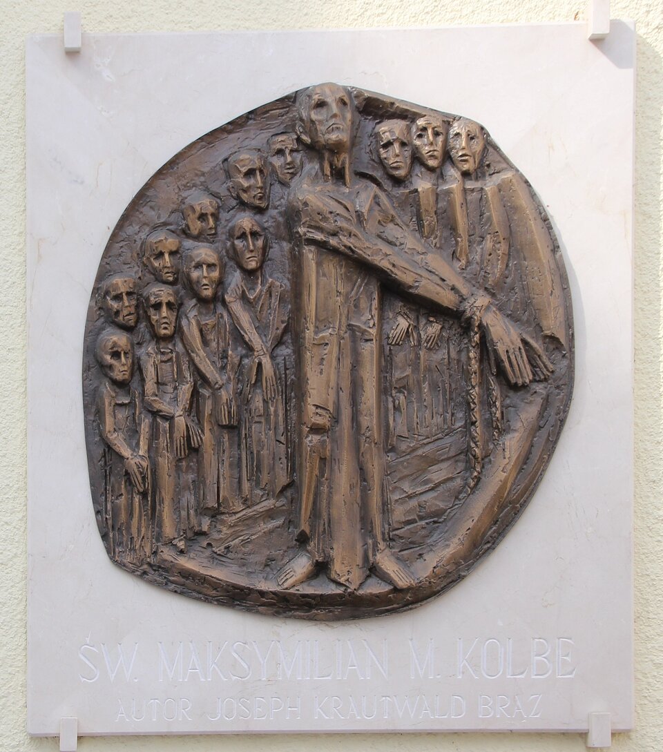 Na zdjęciu tablica z płaskorzeźbą. Płaskorzeźba przedstawia mężczyznę z wyciągniętymi rękoma, które są skute łańcuchem. Za nim znajduje się grupa ludzi. Ich ręce również są skute łańcuchami. Na tablicy pod płaskorzeźbą jest napis Święty Maksymilian Maria Kolbe. Autor Joseph Krautwald. Brąz. 