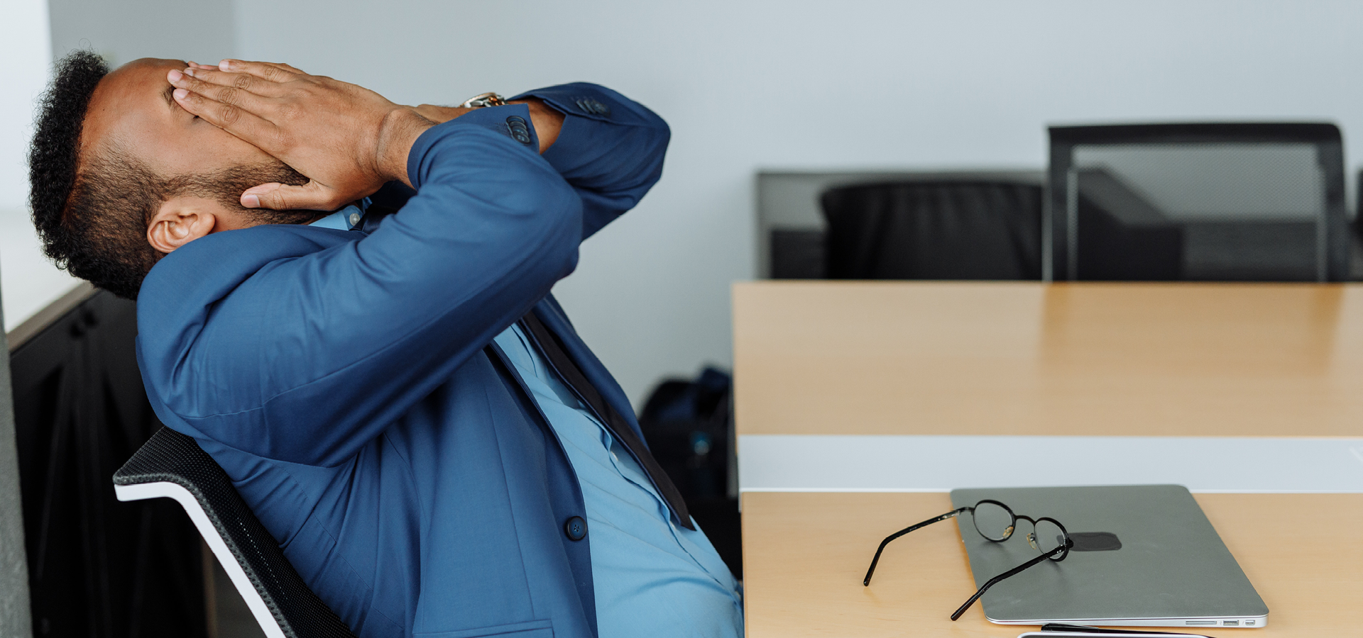 Zdjęcie przedstawia mężczyznę, który siedzi przy biurku. Delikatnie odchyla się na krześle i zasłania twarz dwoma rękami.