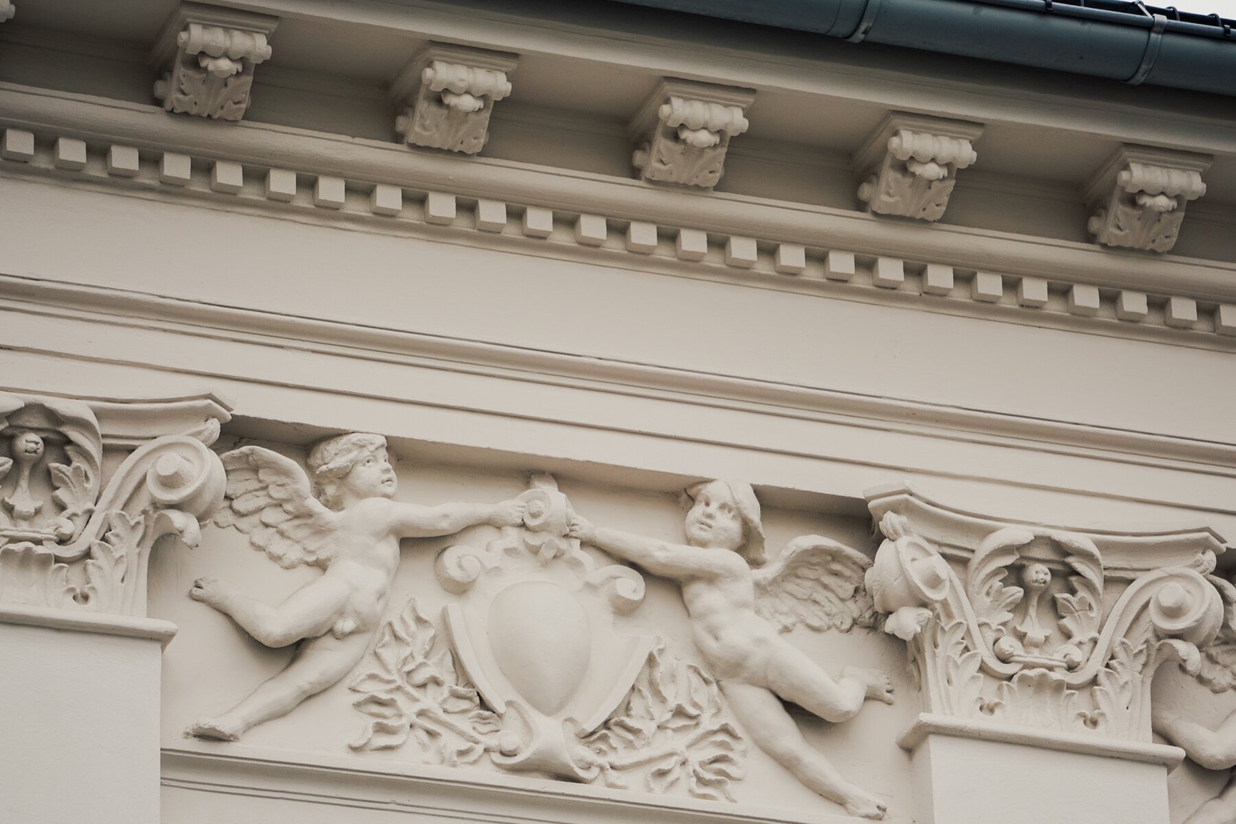Zdjęcie przedstawia gzyms z białego kamienia, na którym znajduje się rzeźba. Przedstawia ona dwa aniołki, które wystawiają rękę do siebie i chwytają za zwijany element dekoracyjny, znajdujący się pomiędzy nimi.