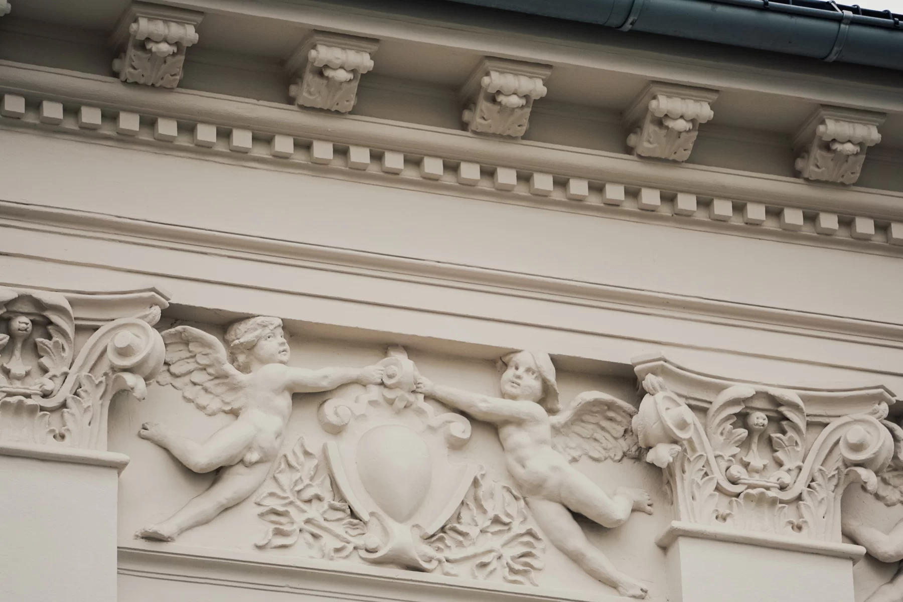 Zdjęcie przedstawia gzyms z białego kamienia, na którym znajduje się rzeźba. Przedstawia ona dwa aniołki, które wystawiają rękę do siebie i chwytają za zwijany element dekoracyjny, znajdujący się pomiędzy nimi.
