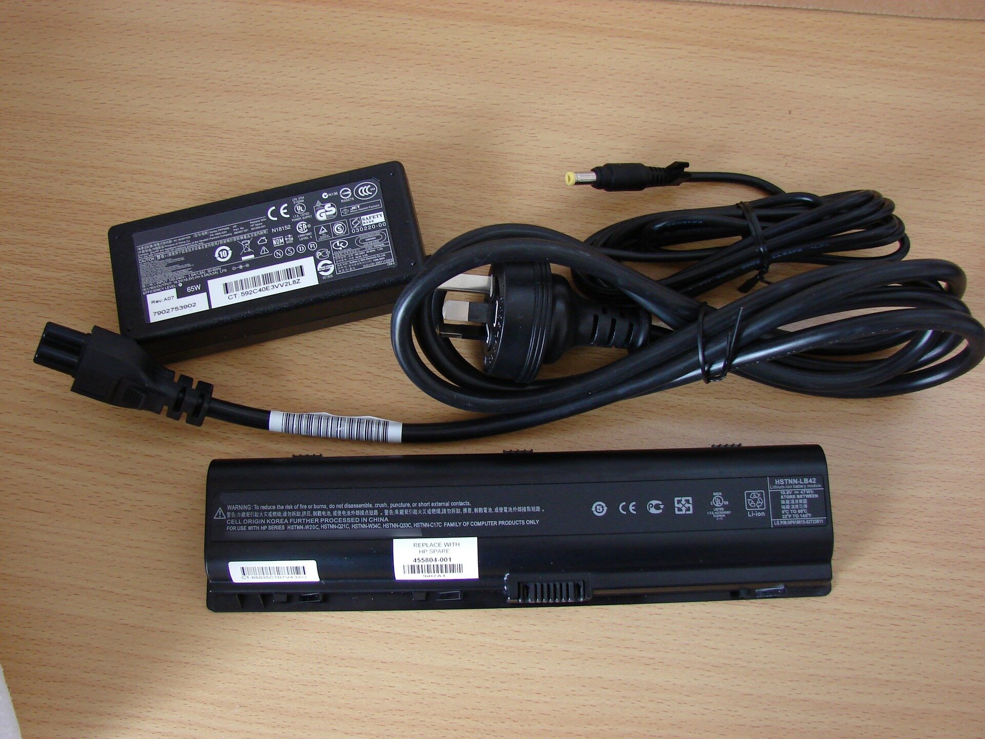 Zdjęcie przedstawia czarny zasilacz do komputera przenośnego w kształcie prostopadłościanu leżący na blacie beżowego biurka wraz z czarnym okablowaniem i akumulatorem do notebooka. Z tabliczki znamionowej zasilacza można odczytać, że parametry prądu na wyjściu to 18,5 wolta i 3,5 ampera.