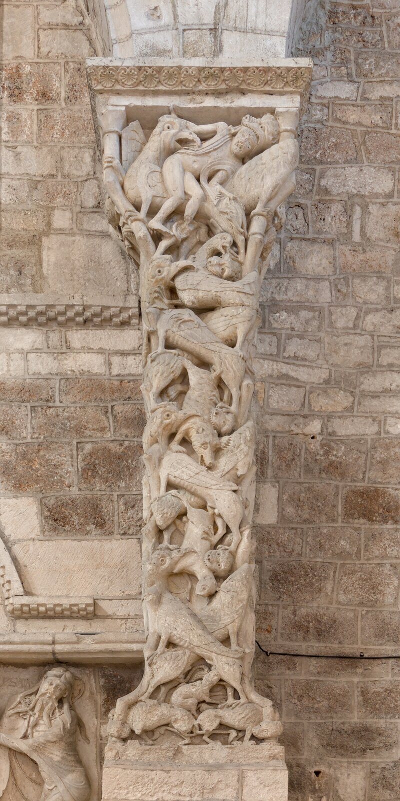 Romańska kolumna rzeźbiona w jasnym kamieniu, gęsto pokryta fantastycznymi zwierzętami i splątanymi postaciami walczącymi ze sobą; dynamiczne, spiralne formy tworzą pionową kompozycję na tle kamiennej ściany kościoła.