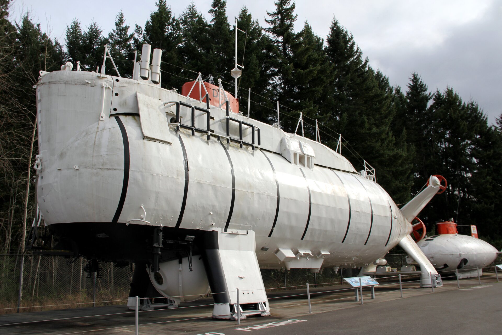 Zdjęcie batyskafu Trieste. Duża, biała, metalowa konstrukcja znajduje się na tle lasu. Ma czerwony kiosk i osiem czarnych pasków namalowanych na boku kadłuba.