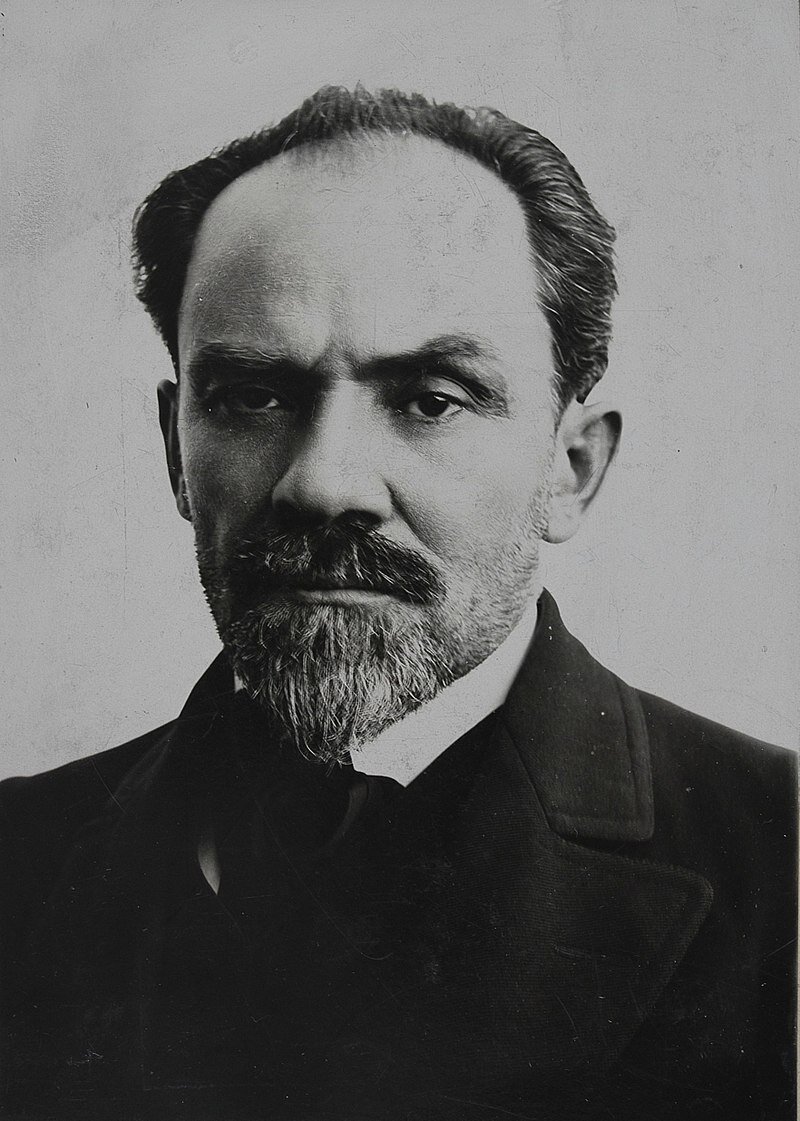 Zdjęcie przedstawia mężczyznę w średnim wieku. Ma wysokie czoło, duży nos, wąsy oraz krótką brodę w szpic. Włosy ma zaczesane do tyłu. Ubrany jest w marynarkę, białą koszulę z kołnierzykiem, krawat.