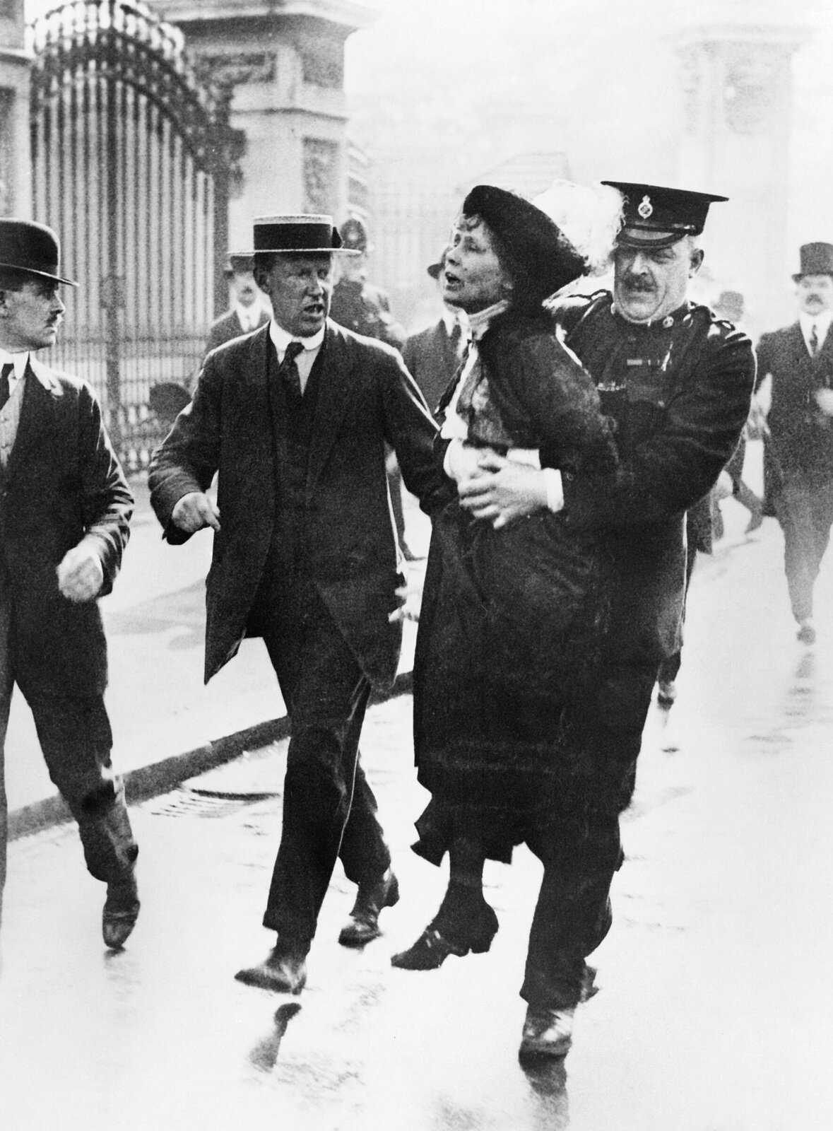 Na zdjęciu ośmiu mężczyzn i jedna kobieta, bohaterka zdjęcia. To Emmeline Pankhurst. Trzyma ją dwóch mężczyzn, jeden z nich w mundurze policyjnym przytrzymuje ja w pasie. W tle metalowa brama Pałacu Buckingham w Londynie.