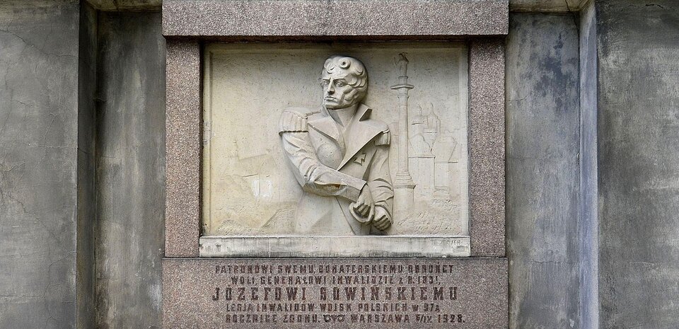Zdjęcie przedstawia tablicę upamiętniającą gen. Józefa Sowińskiego. Składają się na nią płaskorzeźba ostro cięta w jasnym piaskowcu i okalająca ją ciemniejsza rama, złożona z czterech prostokątnych, szarobrązowych kamiennych bloków. Płaskorzeźba ukazuje od bioder w górę postać generała w płaszczu. Głowa ukazana jest lewym półprofilem. Mężczyzna sięga prawą ręką na lewą stronę ciała po szablę, której pochwę przytrzymuje przy lewym biodrze dłoń drugiej ręki. W tle płaskorzeźbiona, jakby zamglona, panorama Warszawy. Na prawo od postaci generała kolumna Zygmunta, a za nią zabudowa miejska. Pod tablicą na płycie ramy napis o treści: PATRONOWI SWEMU BOHATERSKIEMU OBROŃCY WOLI GENERAŁOWI INWALIDZIE Z R. 1831. JÓZEFOWI SOWIŃSKIEMU LEGIA INWALIDÓW WOJSK POLSKICH W 97 Ą ROCZNICĘ ZGONU WARSZAWA 5/IX 1928.