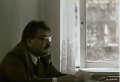 Zdjęcie przedstawia dojrzałego mężczyznę w garniturze, który siedzi przy stoliku i trzyma kartkę. Ma wąsy, krótkie włosy i okulary w dużych oprawkach. Na stole jest telefon. Za mężczyzną jest firanka i lekko otwarte okno.