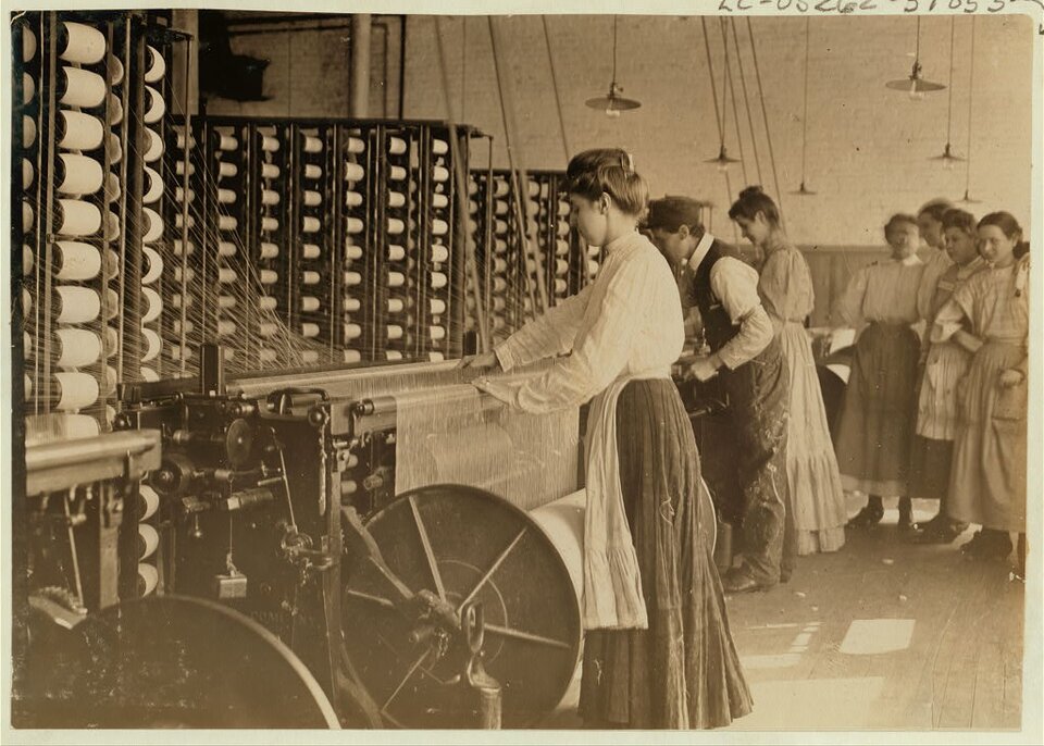 Zdjęcie przedstawia wnętrze XIX- lub wczesno XX‑wiecznej fabryki włókienniczej. W centrum obrazu widać młodą kobietę obsługującą dużą maszynę przędzalniczą. W tle stoi grupa innych pracowników – większość to kobiety, ubrane w długie spódnice i koszule z długimi rękawami, część z nich przygląda się pracy, jaką wykonuje kobieta oraz pozostali pracownicy. Maszyny są duże, mechaniczne, z licznymi szpulami nici i kołami napędowymi. Pomieszczenie jest wysokie, oświetlone elektrycznymi lampami sufitowymi. Maszyny posiadają długie rolki, na które nawijana jest przędza ze szpul umieszczonych na pionowych regałach za maszynami. 