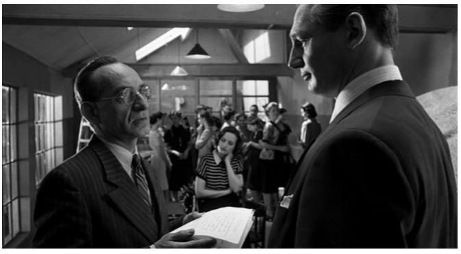 Zdjęcie przedstawia wnętrze fabryki. Na pierwszym planie stoi mężczyzna w garniturze i okularach z kartką w dłoni. Naprzeciw niego stoi Schindler. Na drugim planie znajduje się grupa kobiet. Kobiety stoją, rozmawiają.