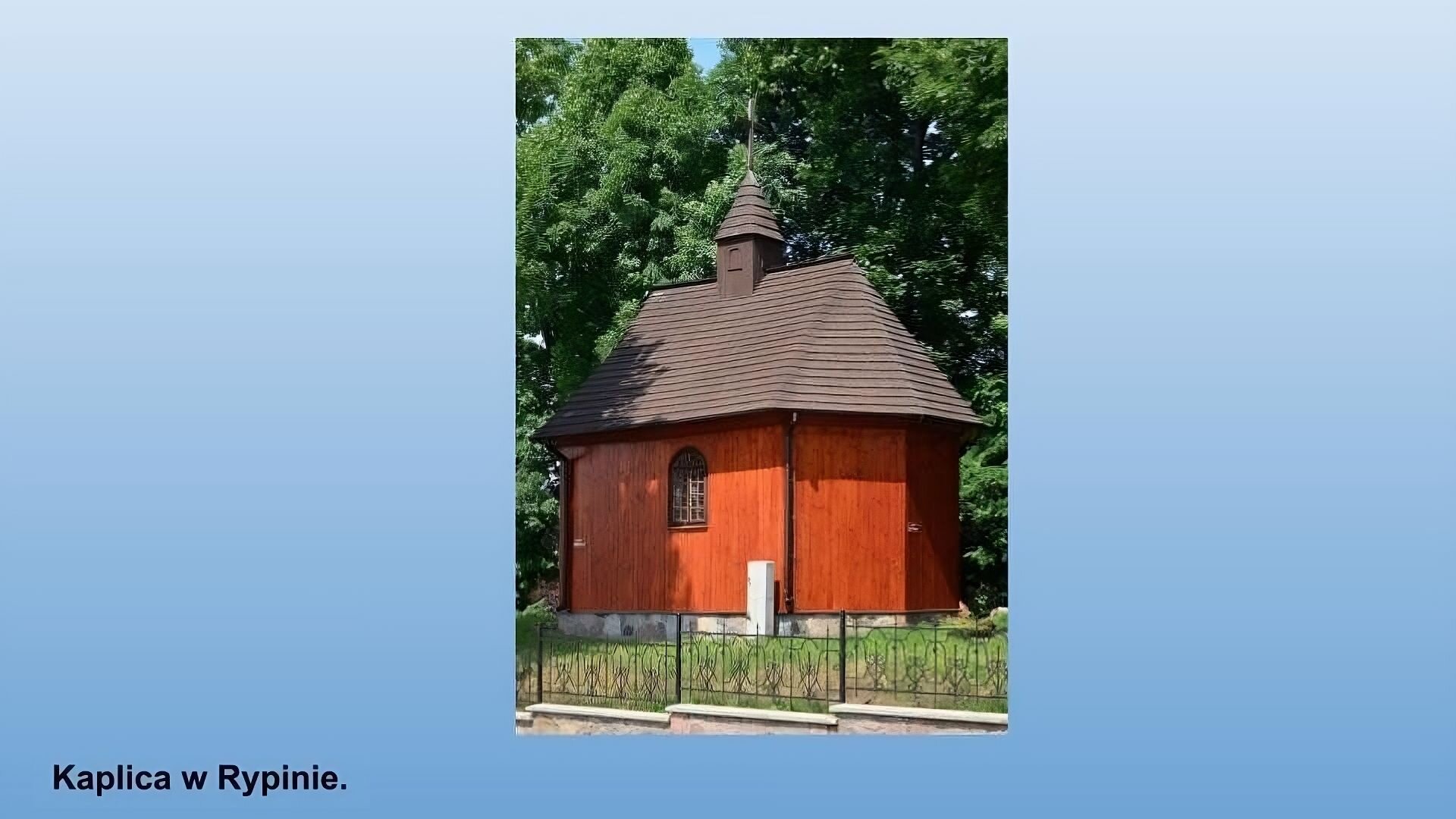 Niebieskie tło. Po środku zdjęcie przedstawiające Kaplicę w Rypinie. Mały budynek o kamiennej podstawie, ścianach z pionowo ułożonych ciemnych desek, z małym oknem z łukiem, z brązowym spadzistym dachem z małą wieżyczką po środku. Przed budynkiem zielona trawa i kuty płotek. W tle drzewa. 