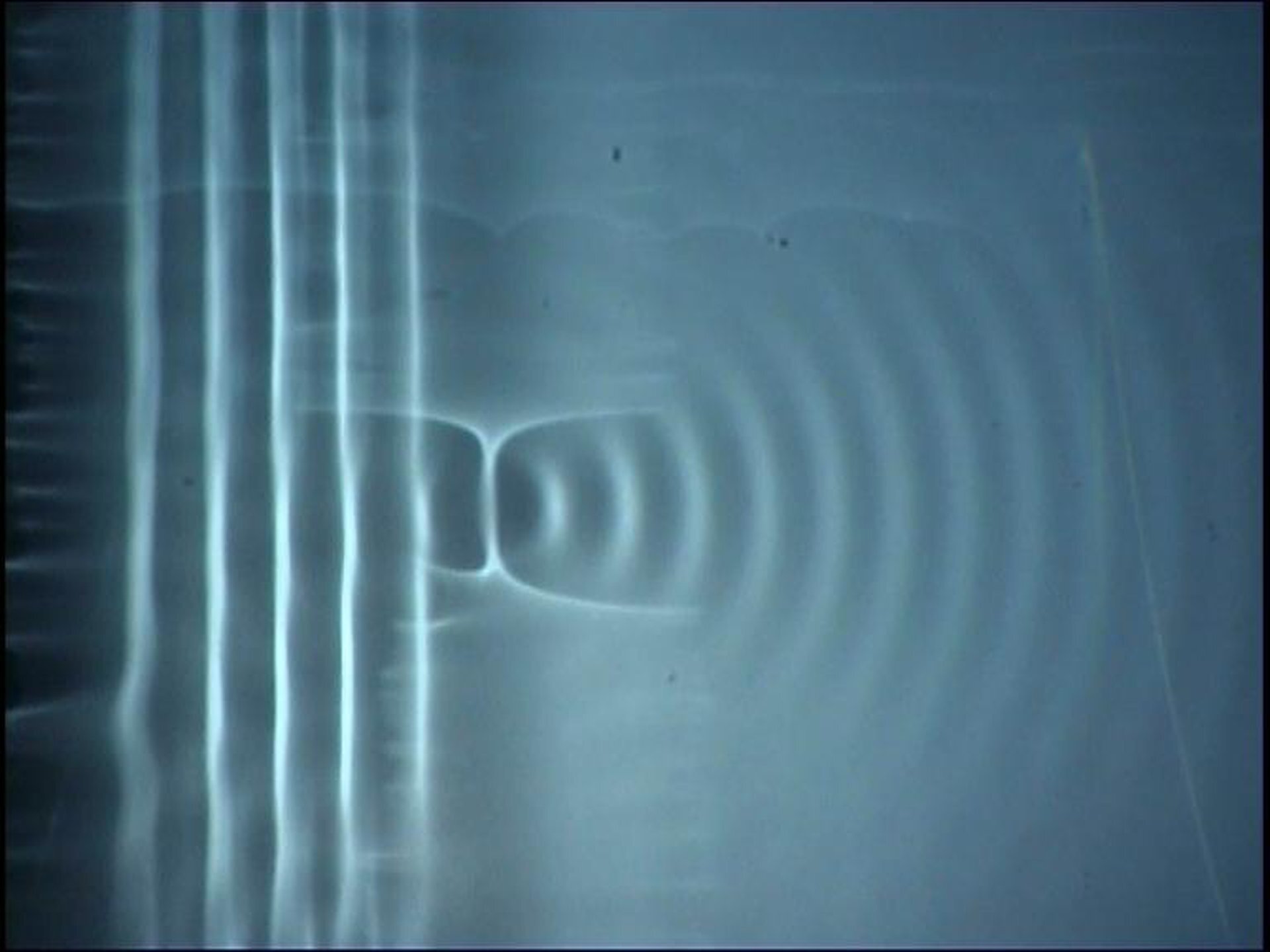 Rys. a. Zdjęcie przedstawia, w jaki sposób fala płaska jest wytwarzana na powierzchni wody za pomocą drgającej deski. Kuweta z wodą sfotografowana jest od góry. Po lewej stronie widać obszar, gdzie rozprzestrzenia się front falowy. Są to pionowe, równoległe prążki, które powstały w wyniku uderzenia deski w wodę. Na środku znajduje się przeszkoda. Po jej prawej stronie fala rozchodzi się już koliście. Tworzy współśrodkowe półokręgi. 