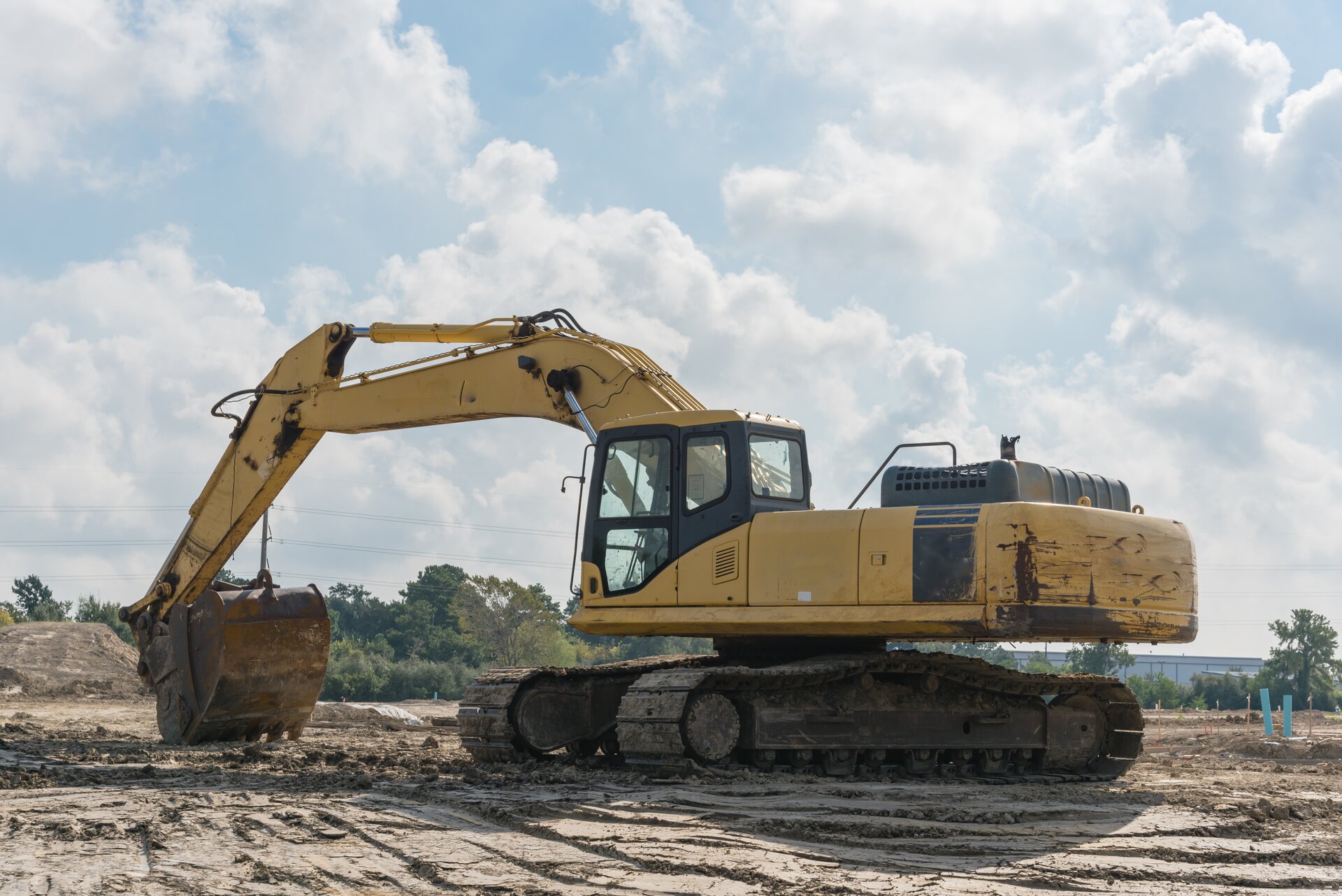 Zdjęcie przedstawia koparkę przedsiębierną. Widoczna jest żółta koparka, w ujęciu z boku, stojąca na rozjeżdżonej ziemi. Ramię koparki jest wyprostowane, a łyżka jest podniesiona nad ziemią. Po lewej stronie od ramienia widoczna jest kabina operatora koparki, a na jej tyle duża pozioma platforma. W tle widoczną hałdy ziemi, plac budowy, a na horyzoncie drzewa.

Opisy obiektów prezentowanych na grafikach są załączone w treści galerii zdjęć.