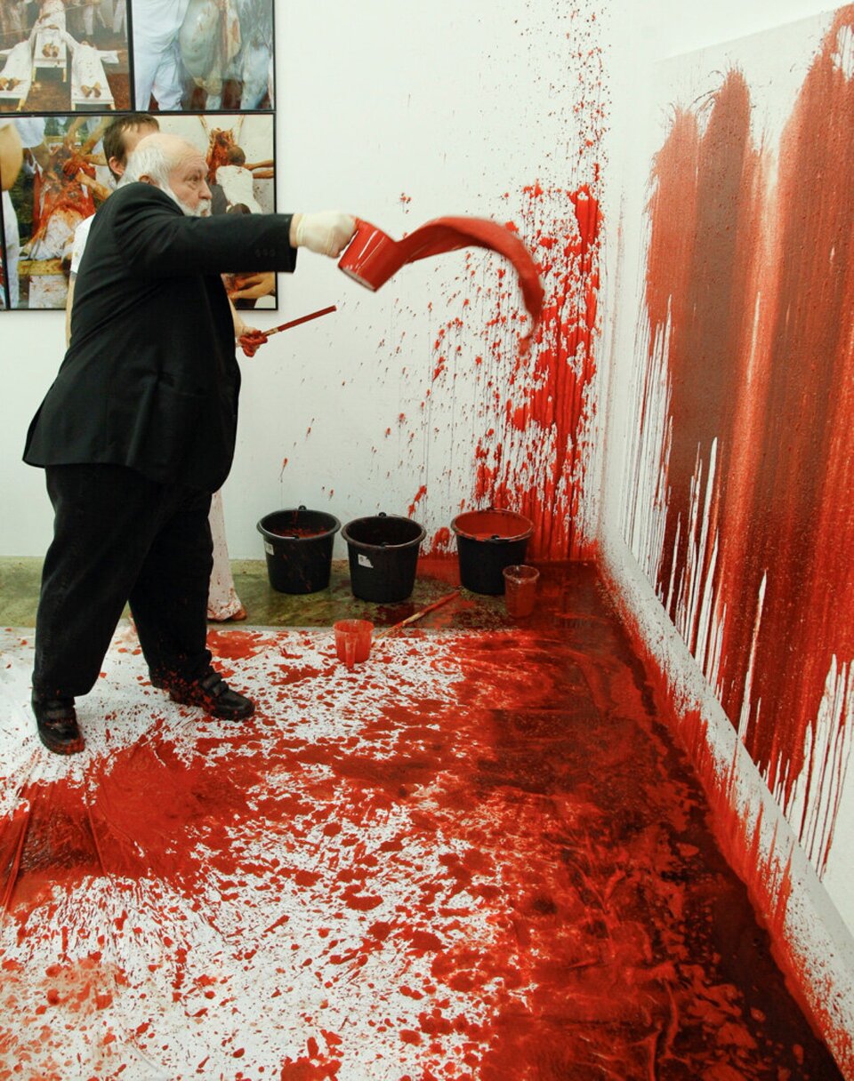 Fotografia przedstawia starszego, siwowłosego mężczyznę w trakcie performance’u. Mężczyzna jest niski i korpulentny, ubrany w ciemną marynarkę i spodnie. Ma siwą brodę i wąsy. Jego ciemne buty ubrudzone są czerwoną farbą. Mężczyzna znajduje się w pomieszczeniu zachlapanym czerwoną farbą. Farba znajduje się w dużej ilości, zarówno  na ścianach, jak i na podłodze. Farba została rozchlapana z użyciem siły oraz z rozmaitej odległości. Na ścianach przeważają podłużne, spływające smugi, zaś na podłodze plamy różnej wielkości. W prawej ręce mężczyzna trzyma pojemnik z farbą, którą również z rozmachem chlusta o ścianę. Farba została uchwycona w sposób dynamiczny, w locie. Wydostała się już z wiaderka, ale nie dotknęła jeszcze ściany. W kącie pomieszczenia znajdują się pojemniki z czerwoną farbą.  Na ścianie po lewej stronie znajdują się cztery zdjęcia o tematyce sakralnej.