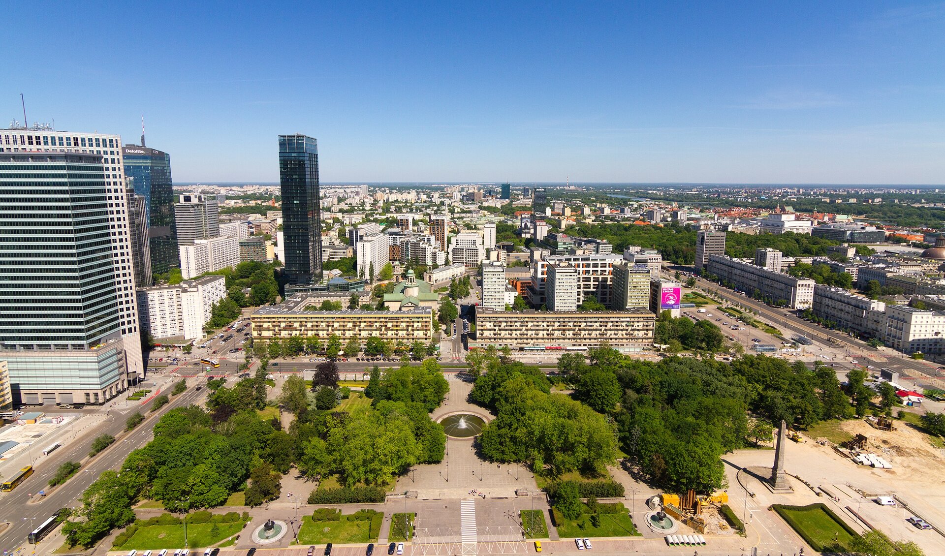Zdjęcie przedstawia panoramę miasta. Na pierwszym planie jest plac, na którym jest fontanna i kilka drzew. Po lewej stronie stoją trzy wysokie, szklane wieżowce. Na wprost i po prawej stronie ciągną się kamienice i bloki.